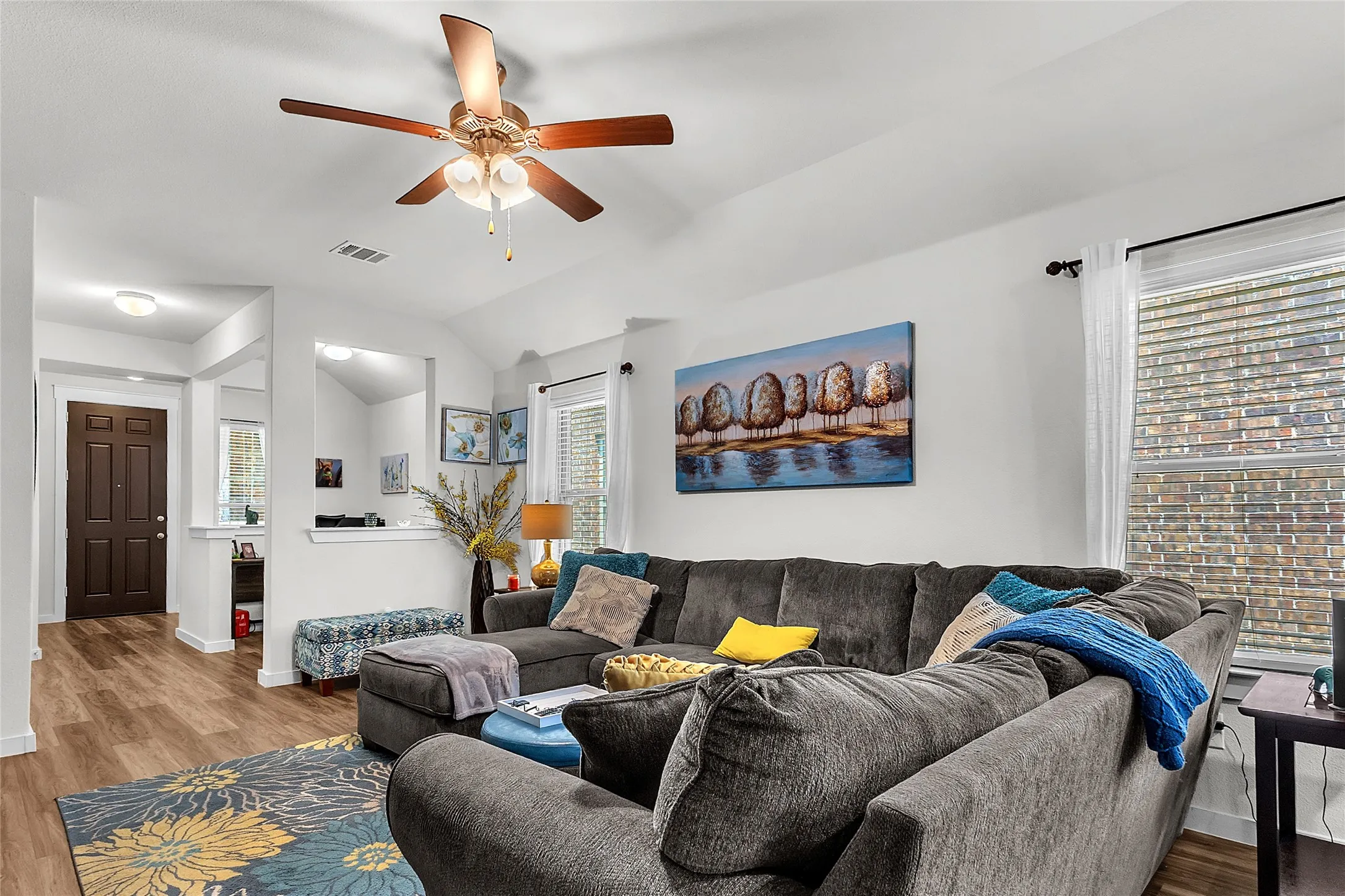 Living room with LVP flooring, gray sectional sofa, blue accent pillows, ceiling fan, and natural light from window with a view of outdoor greenery