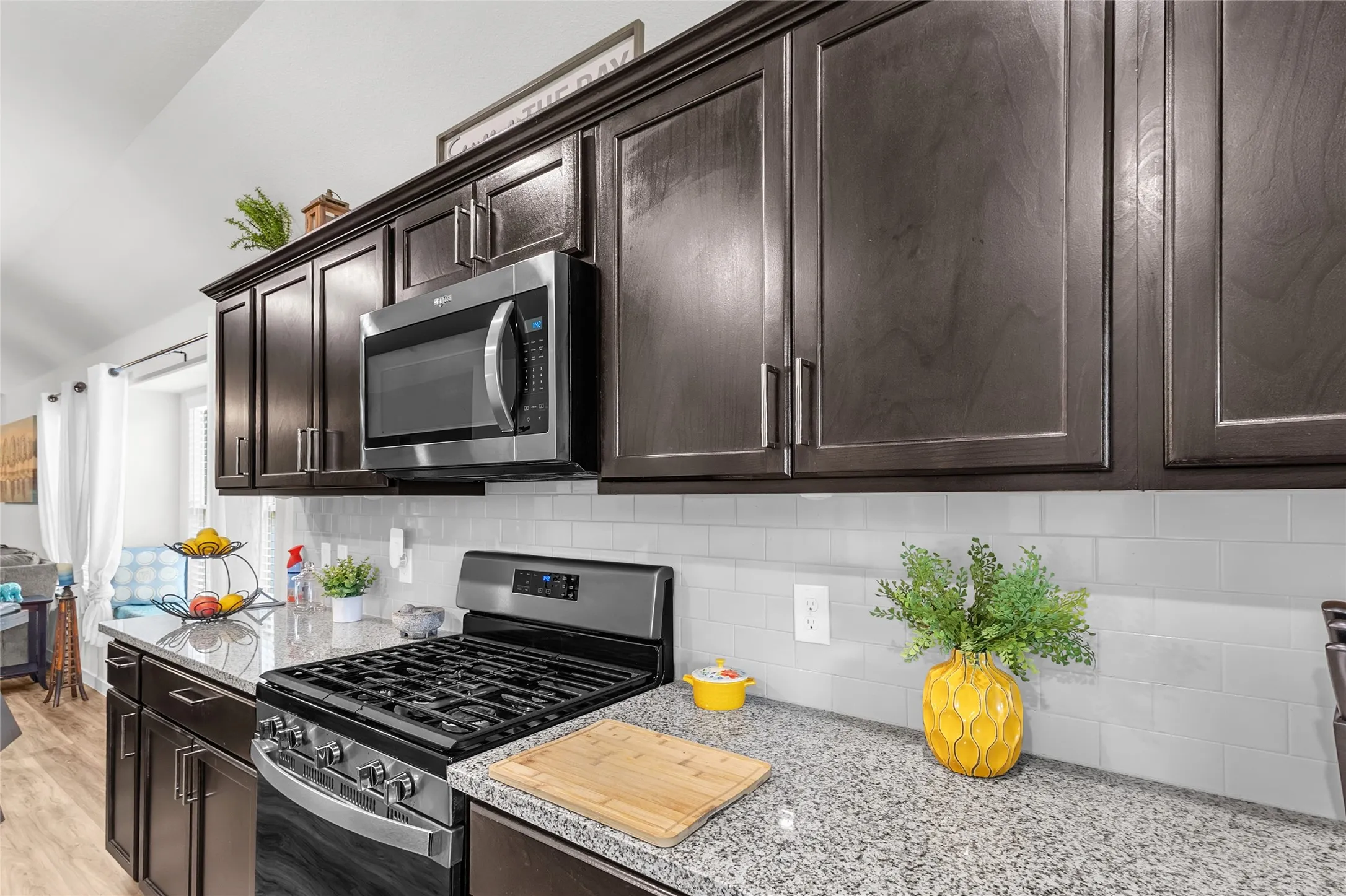 Open-plan kitchen with granite countertops, dark wood cabinetry, stainless steel appliances, and light-colored backsplash accented by natural light from nearby windows