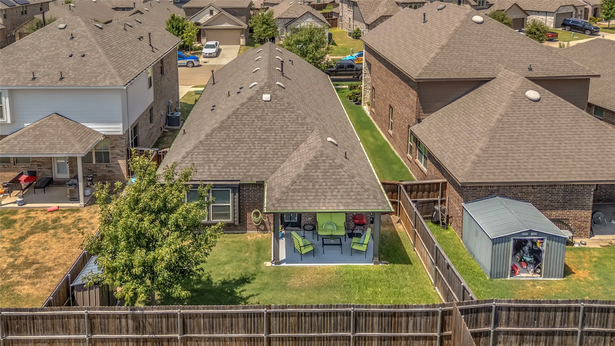 Covered back patio with green seating, adjacent to a well-maintained lawn and neighborhood houses, surrounded by wooden fencing and a storage shed