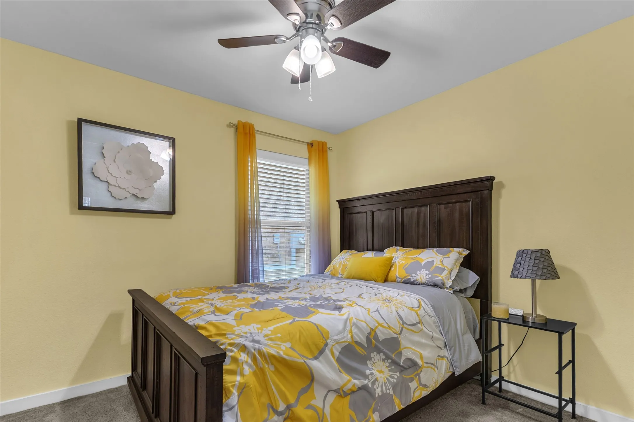 Primary bedroom with a wooden bed frame, yellow floral bedding, and a side table, illuminated by natural light from a window with sheer curtains