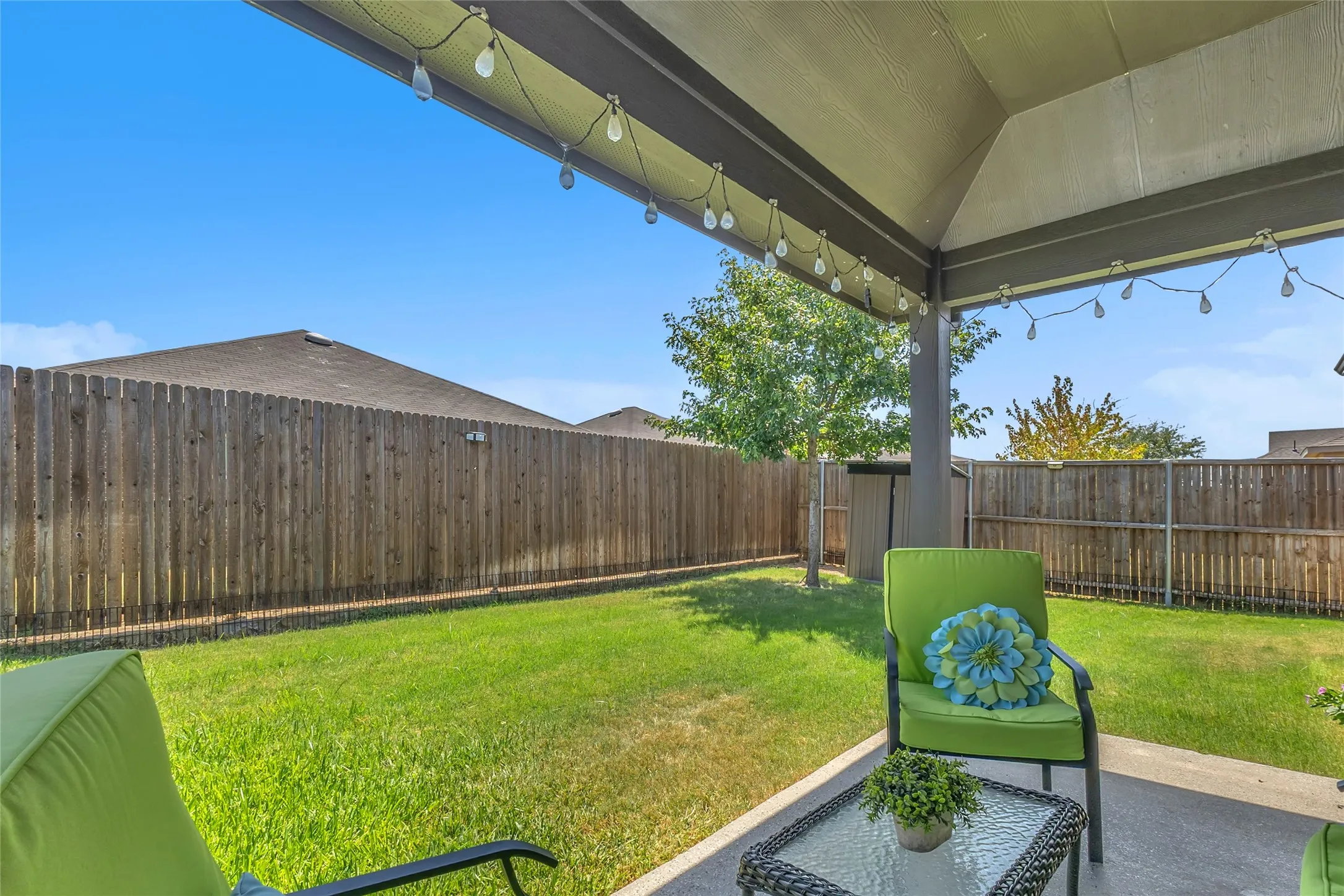 Covered back patio with green lawn, decorative string lights, and two lounge chairs on a concrete surface, surrounded by wooden fencing and trees