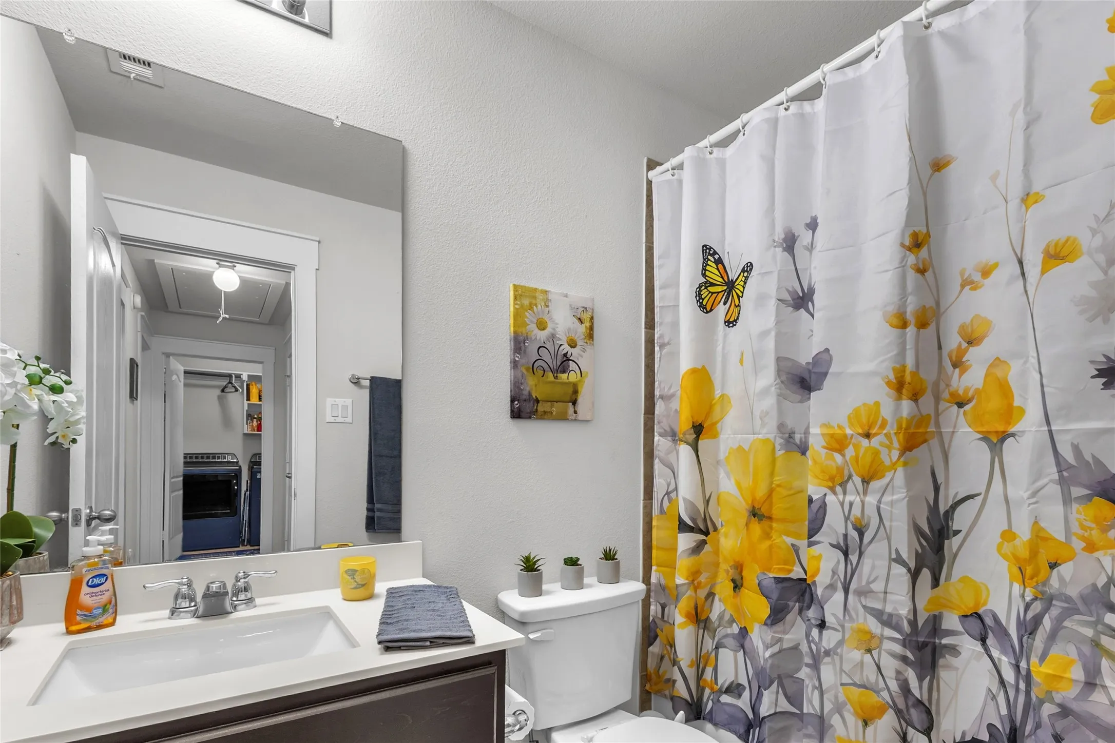 Bathroom with floral shower curtain, rectangular mirror, quartz countertop, undermount sink, and small potted plants under soft overhead lighting