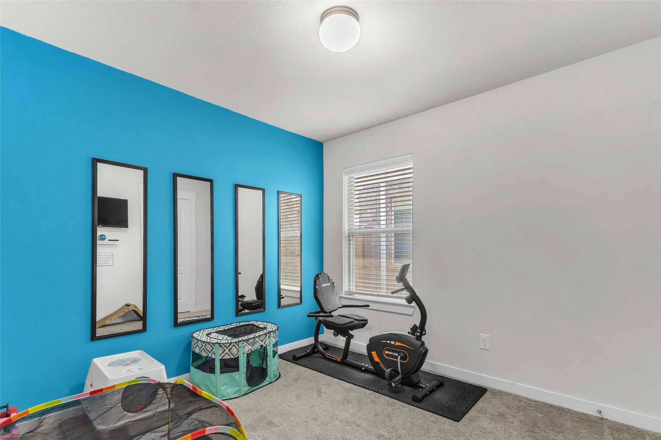 Home gym with an exercise bike, three wall mirrors, textured blue accent wall, and a cozy carpeted floor illuminated by a ceiling light.