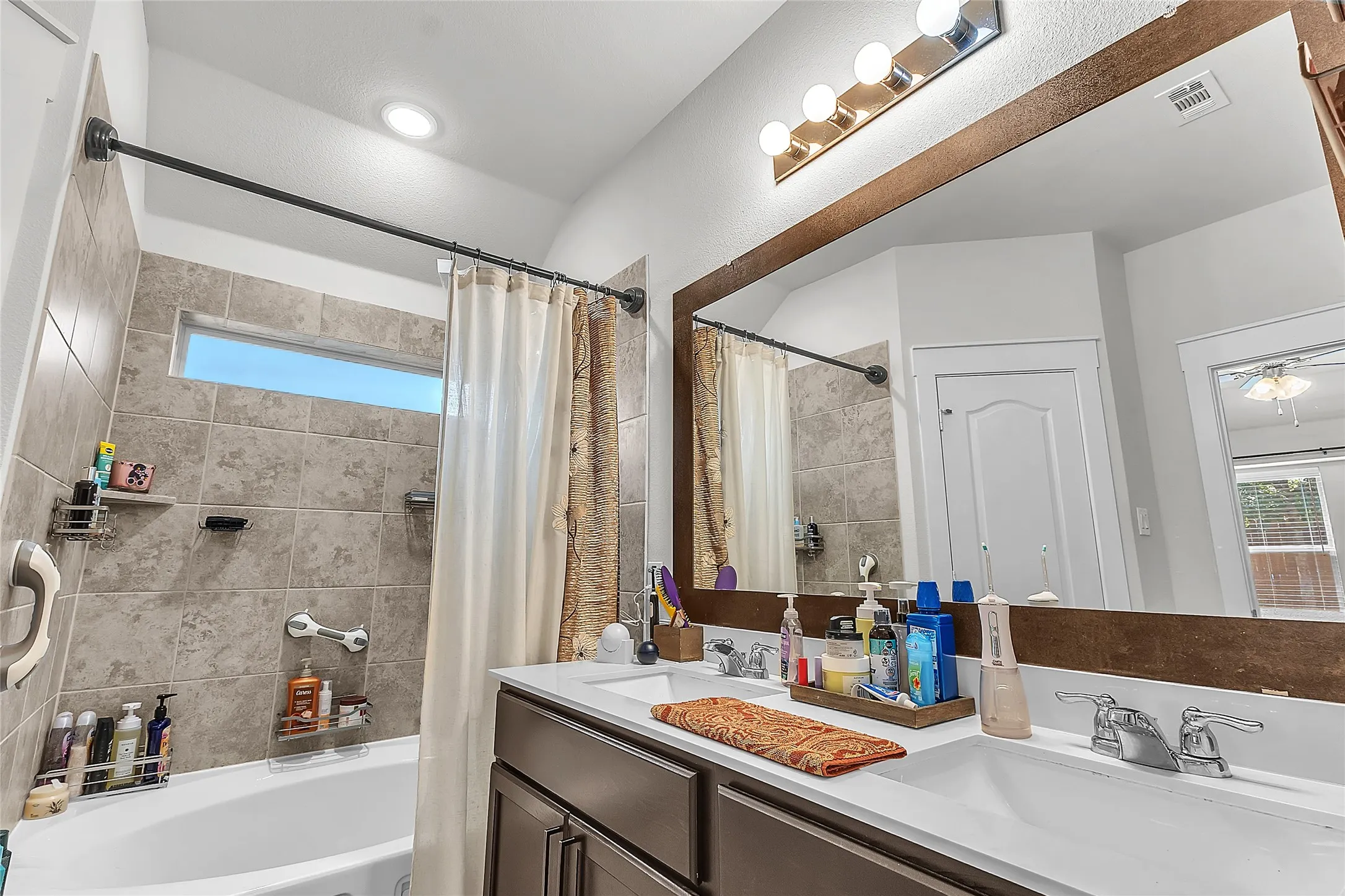 Bathroom with dual sinks, tan tiled walls, light wood accents, a shower/tub combo, natural light from a window, and a large wall mirror.