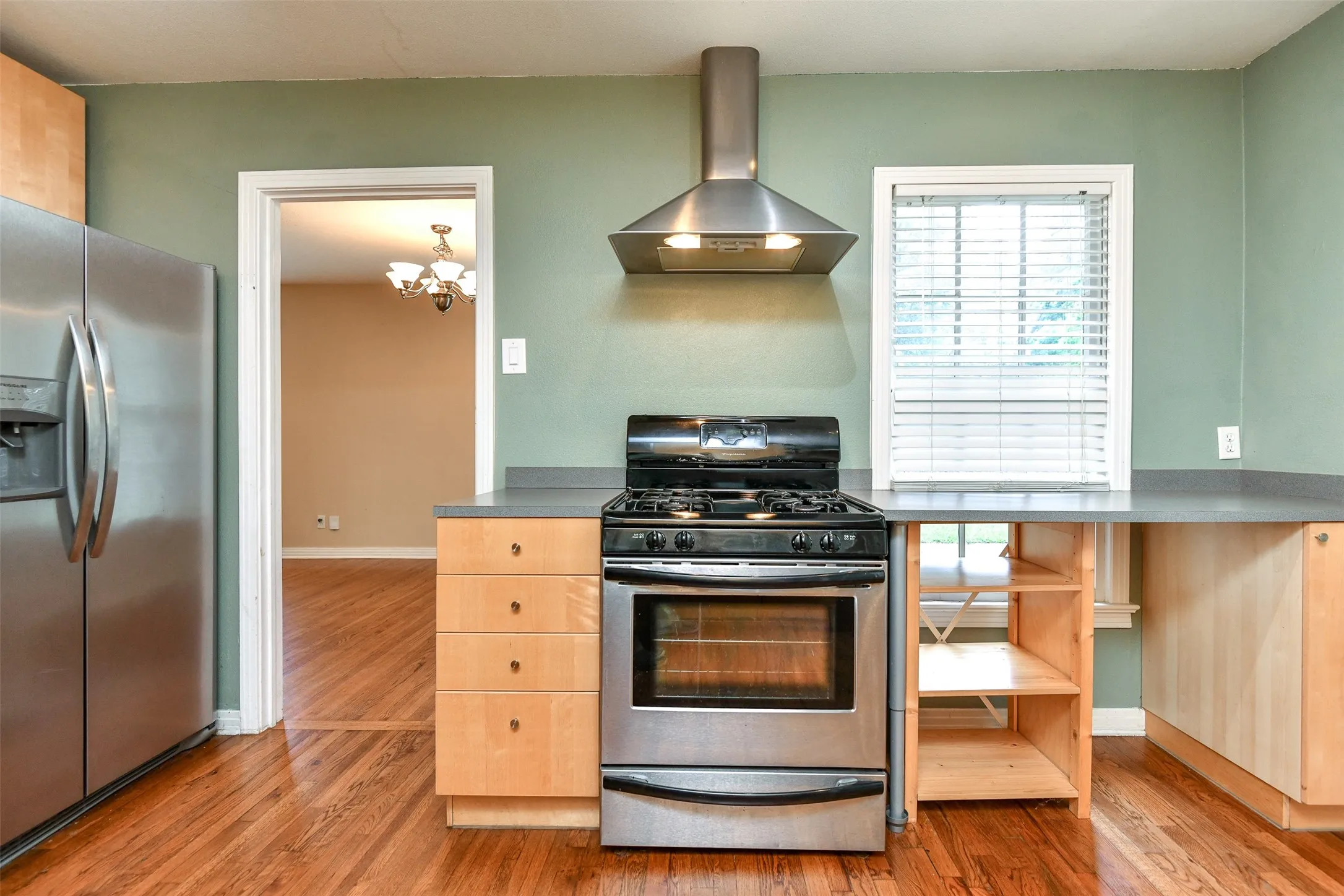 Kitchen featuring appliances with stainless steel finishes, light brown cabinetry, wall chimney range hood, a chandelier, and hardwood / wood-style floors