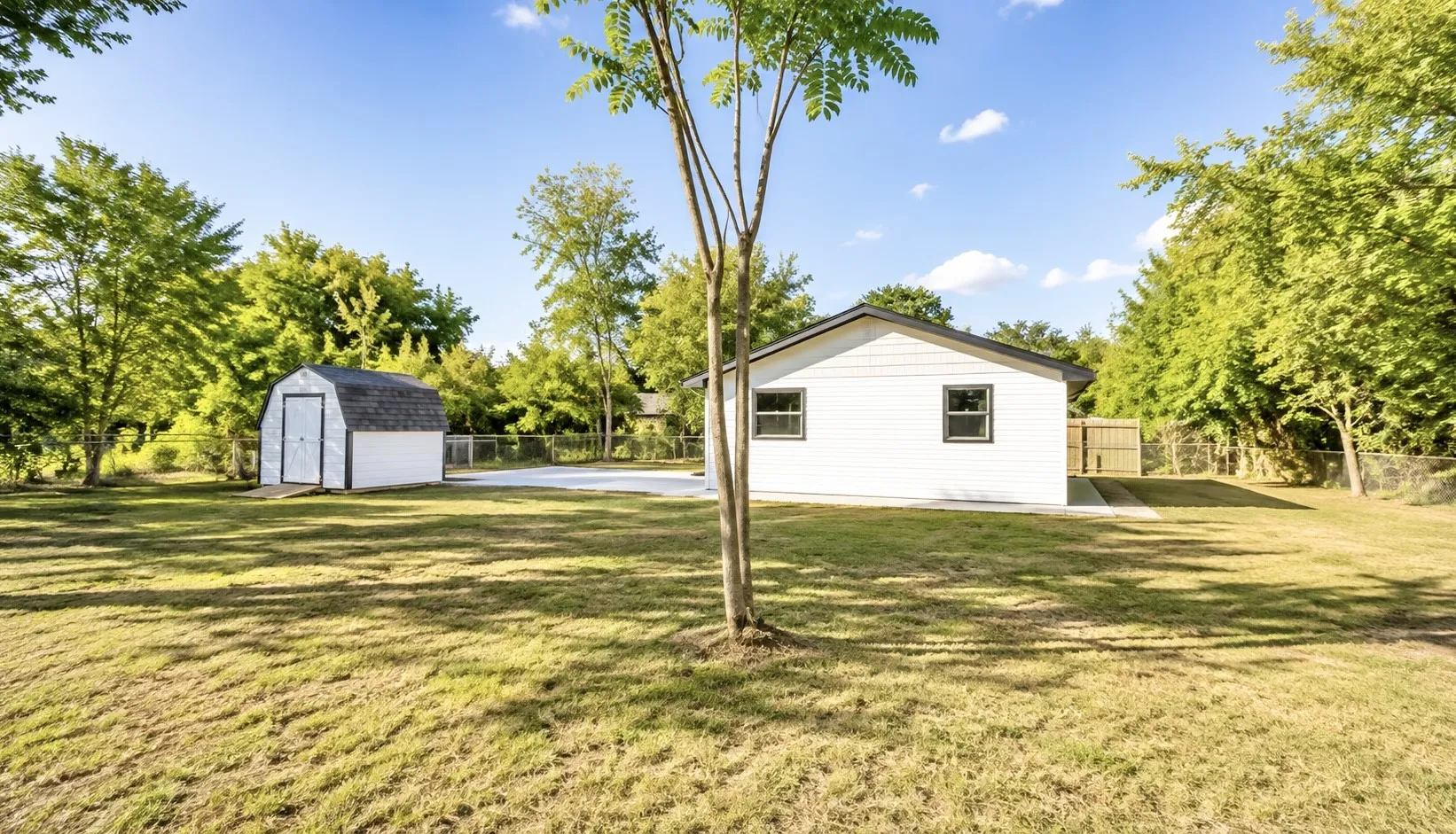 Expansive grassy yard featuring a gabled storage shed, concrete patio, and a chain-link fence
