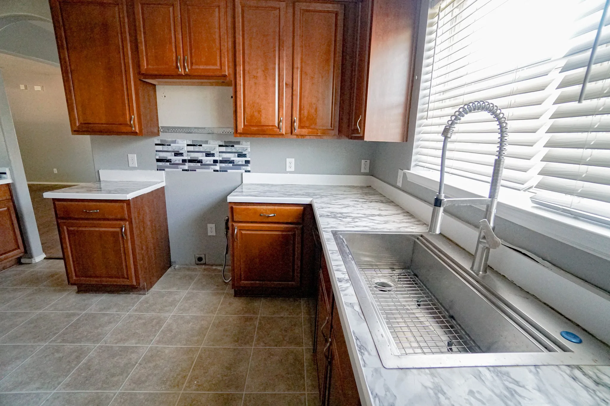 Kitchen featuring wood cabinetry, marbled countertops, a stainless steel sink with a gooseneck faucet, a patterned tile backsplash, and tile flooring