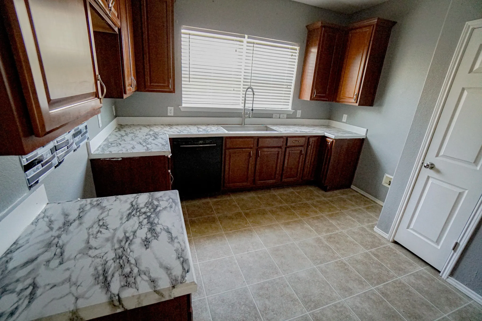 Kitchen featuring wood cabinetry, white-and-gray patterned countertops, a stainless steel sink with a gooseneck faucet, a black dishwasher, and tiled flooring