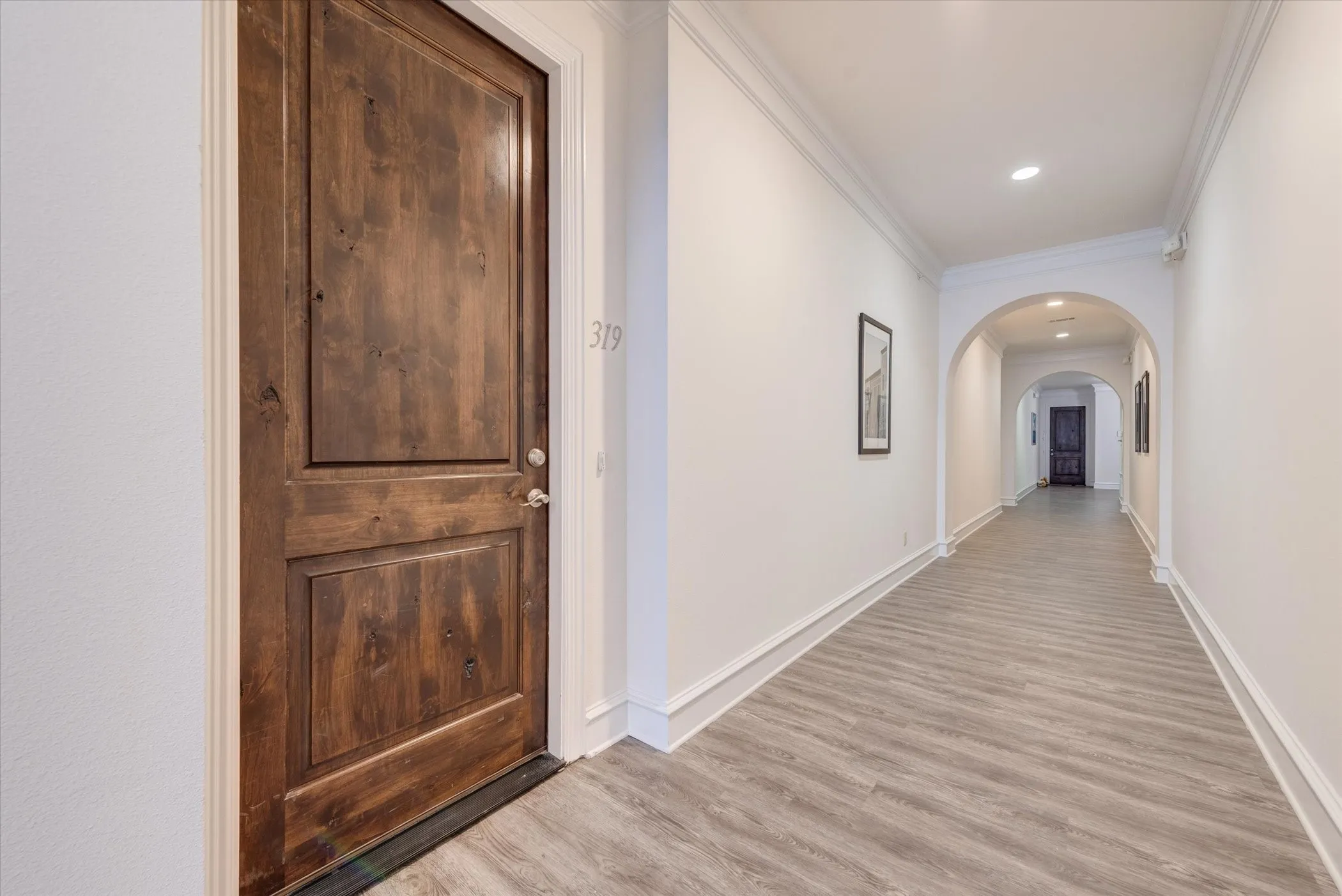 Corridor featuring a rich wood-paneled door, light wood-finish flooring, white crown molding, recessed lighting, and arched doorways