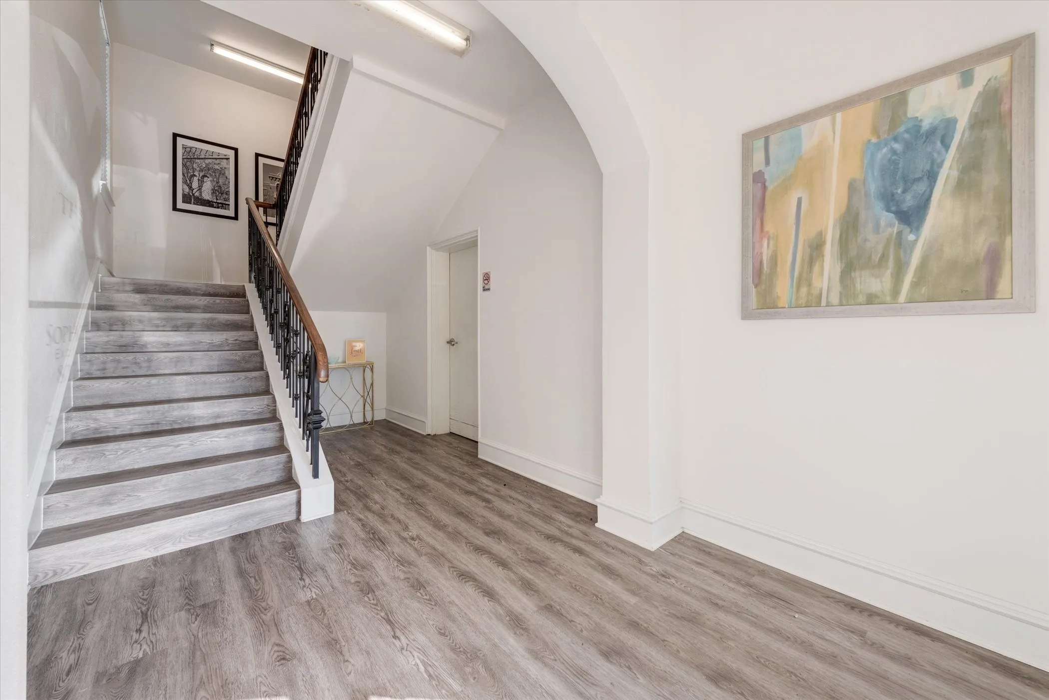 Lobby with wood-finish flooring, staircase with dark metal and wood banister, arched entryway, and white walls