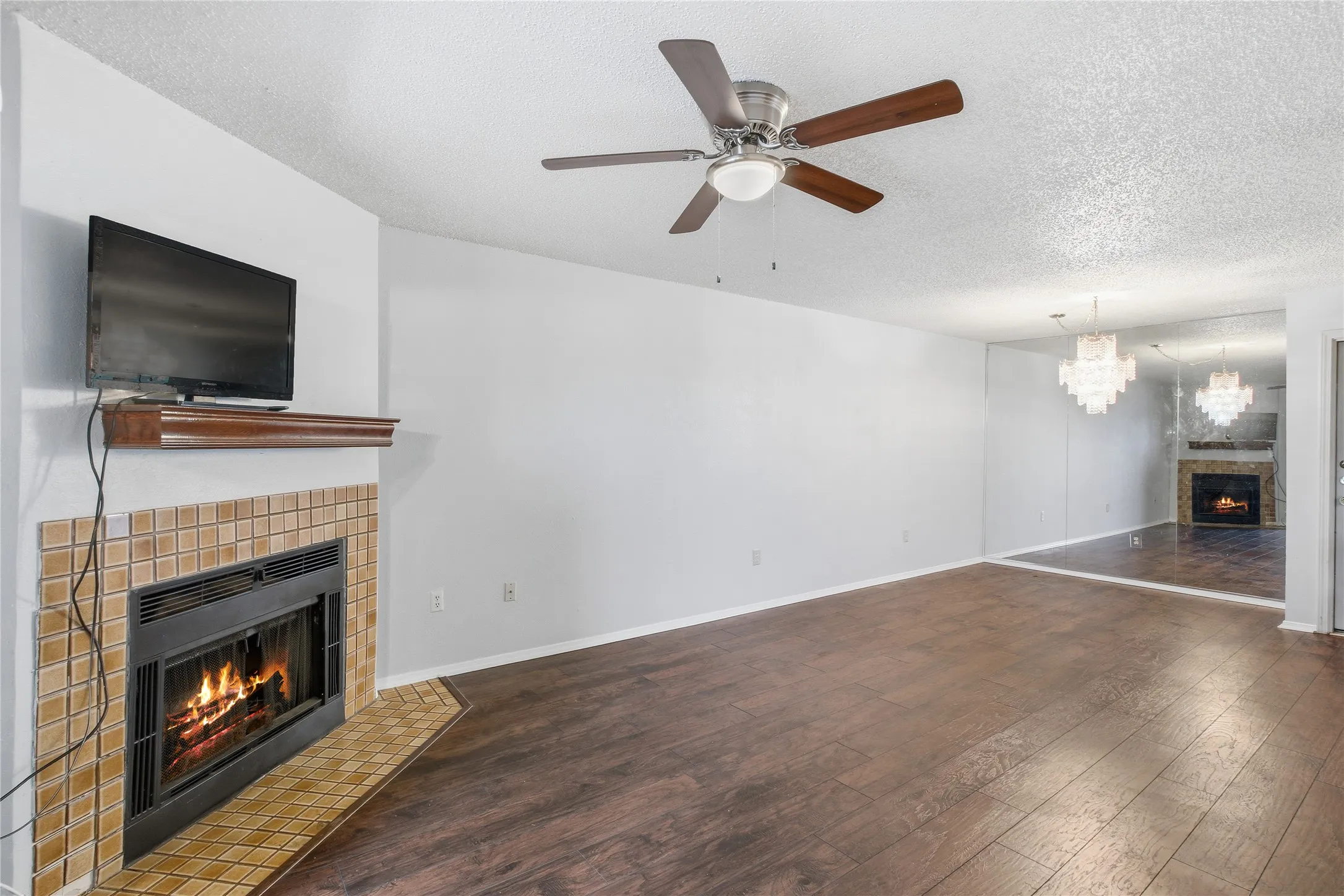 Wood-finish flooring, tiled fireplace surround with hearth, wooden mantel, ceiling fan with integrated lighting, and mirrored wall accent