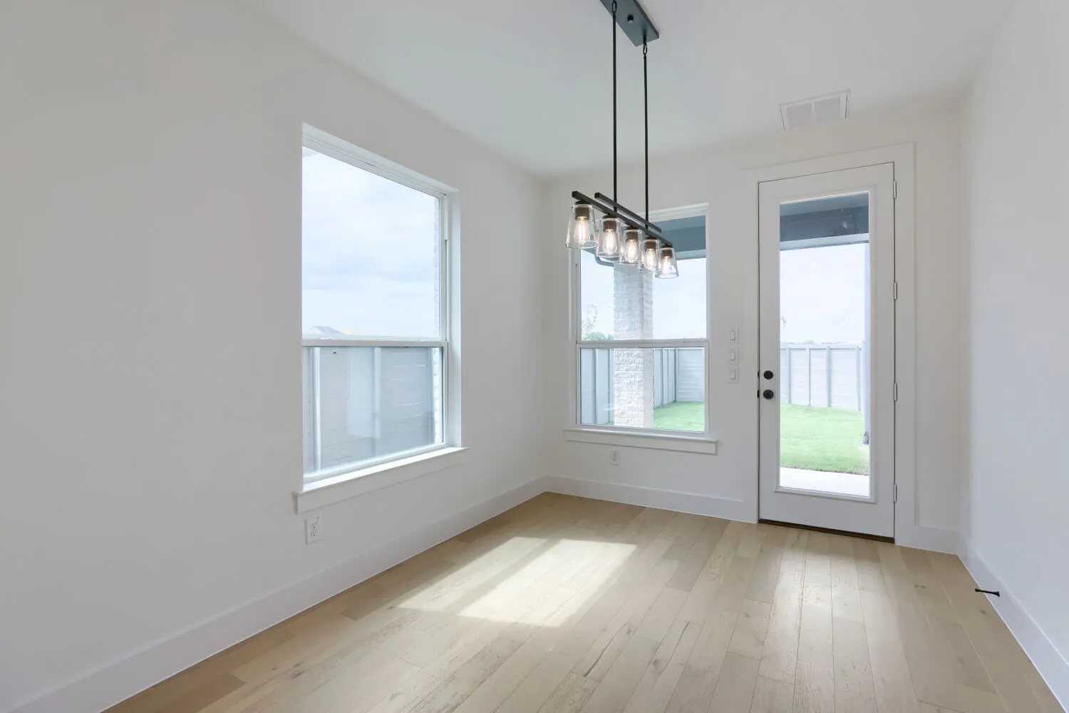 Bright, open living space featuring hardwood flooring, abundant natural light from dual windows and glass door access to outdoor space, and a modern pendant light fixture suspended from the ceiling