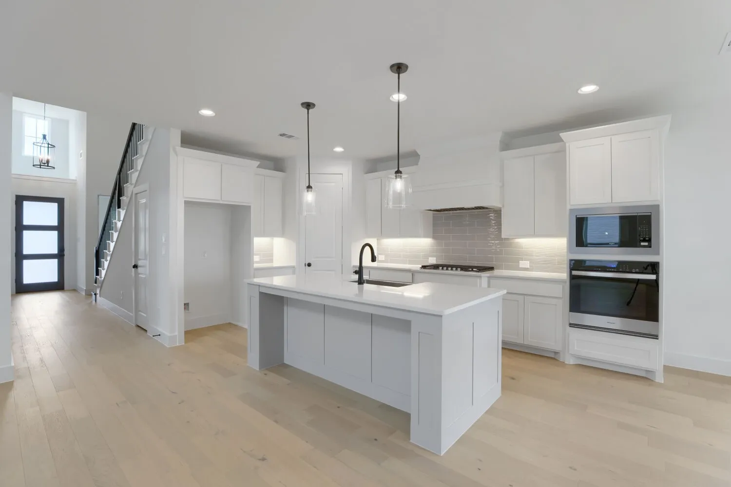 White shaker cabinetry, light wood-finish flooring, stainless steel built-in appliances, pendant lighting, and a kitchen island with a sink