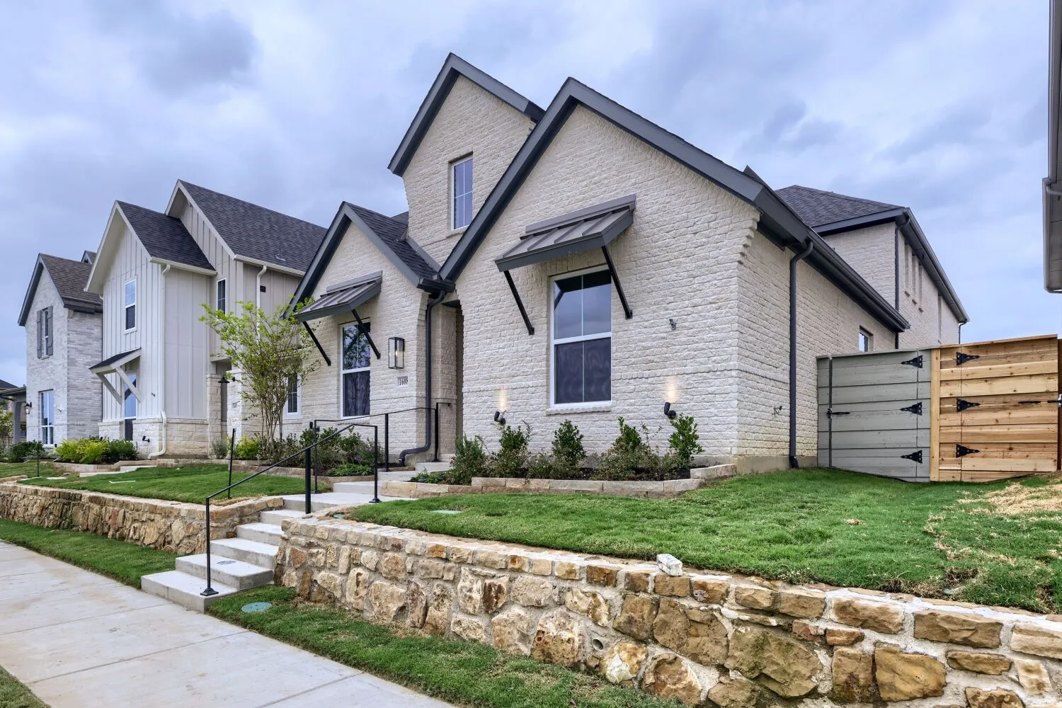 White brick exterior, black trim accents, decorative window awnings, stone retaining wall, and manicured landscaping