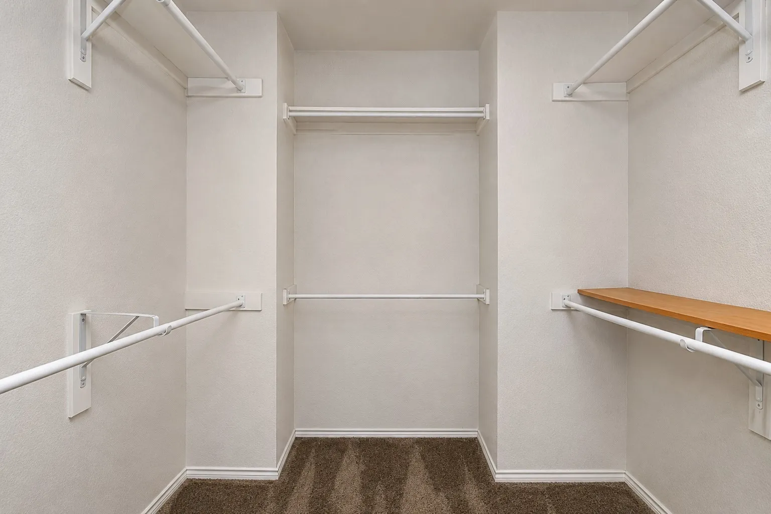 Closet space featuring white walls, carpeted flooring, white hanging rods, and a wood-finish shelf