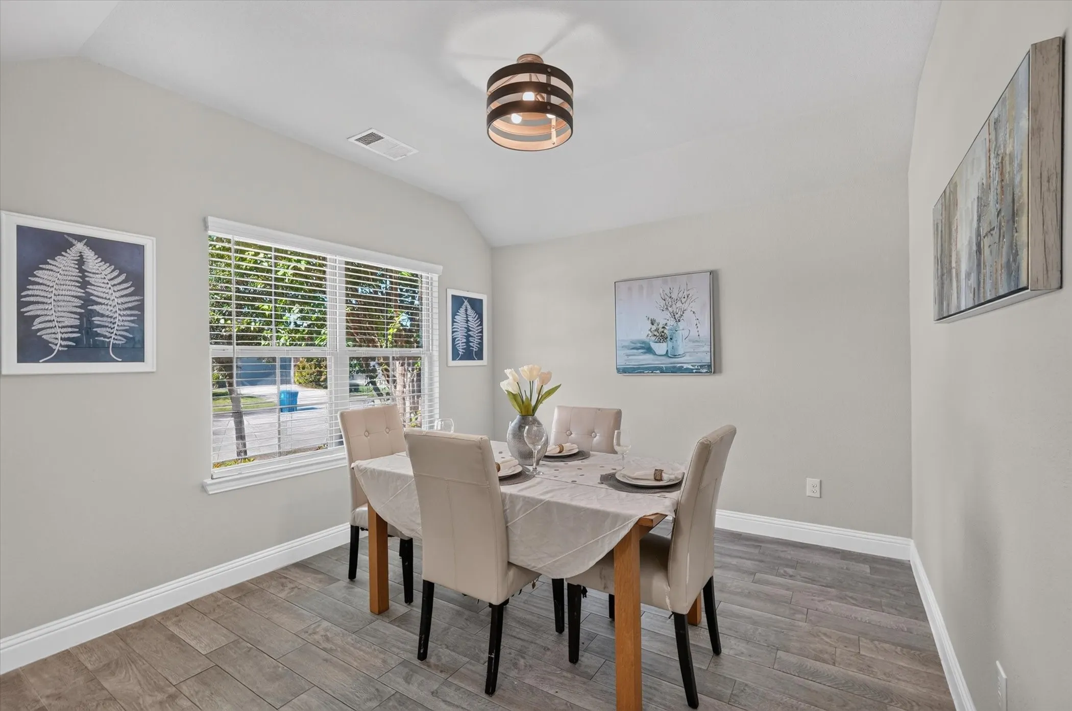 Bright dining room showcasing wood-finish flooring, large multi-pane windows with natural light, neutral wall finishes, a striped drum pendant ceiling fixture, and space for a dining table with seating for multiple guests