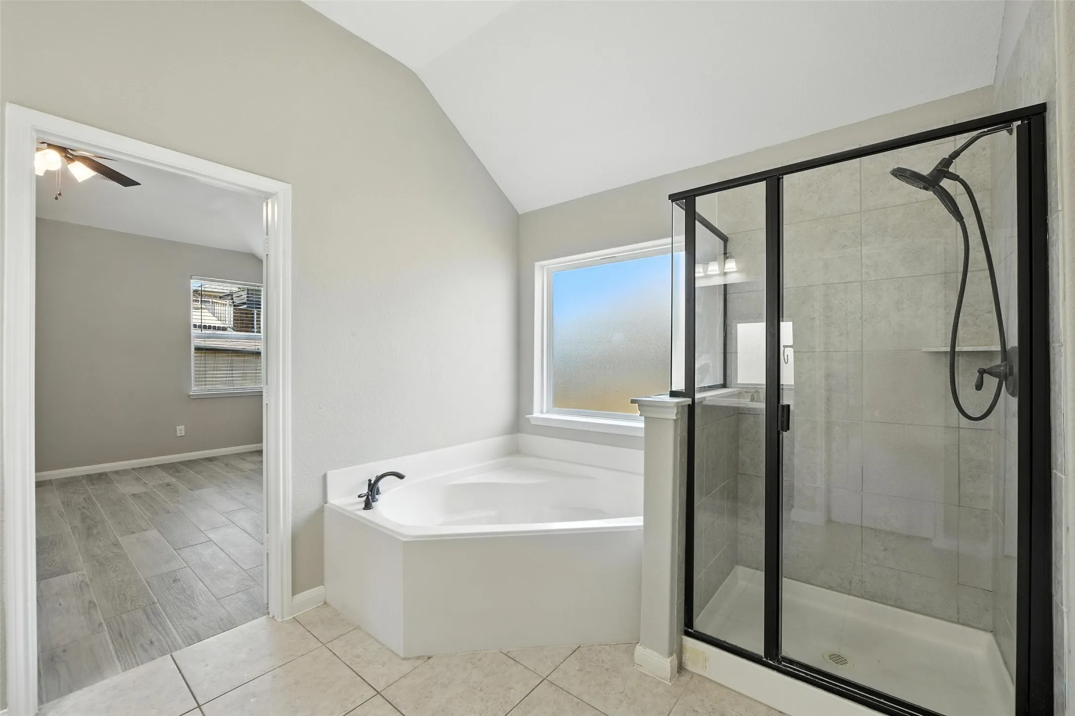 Soaking tub with dark bronze fixtures, glass-enclosed shower with dark bronze framing, vaulted ceiling, and tile flooring