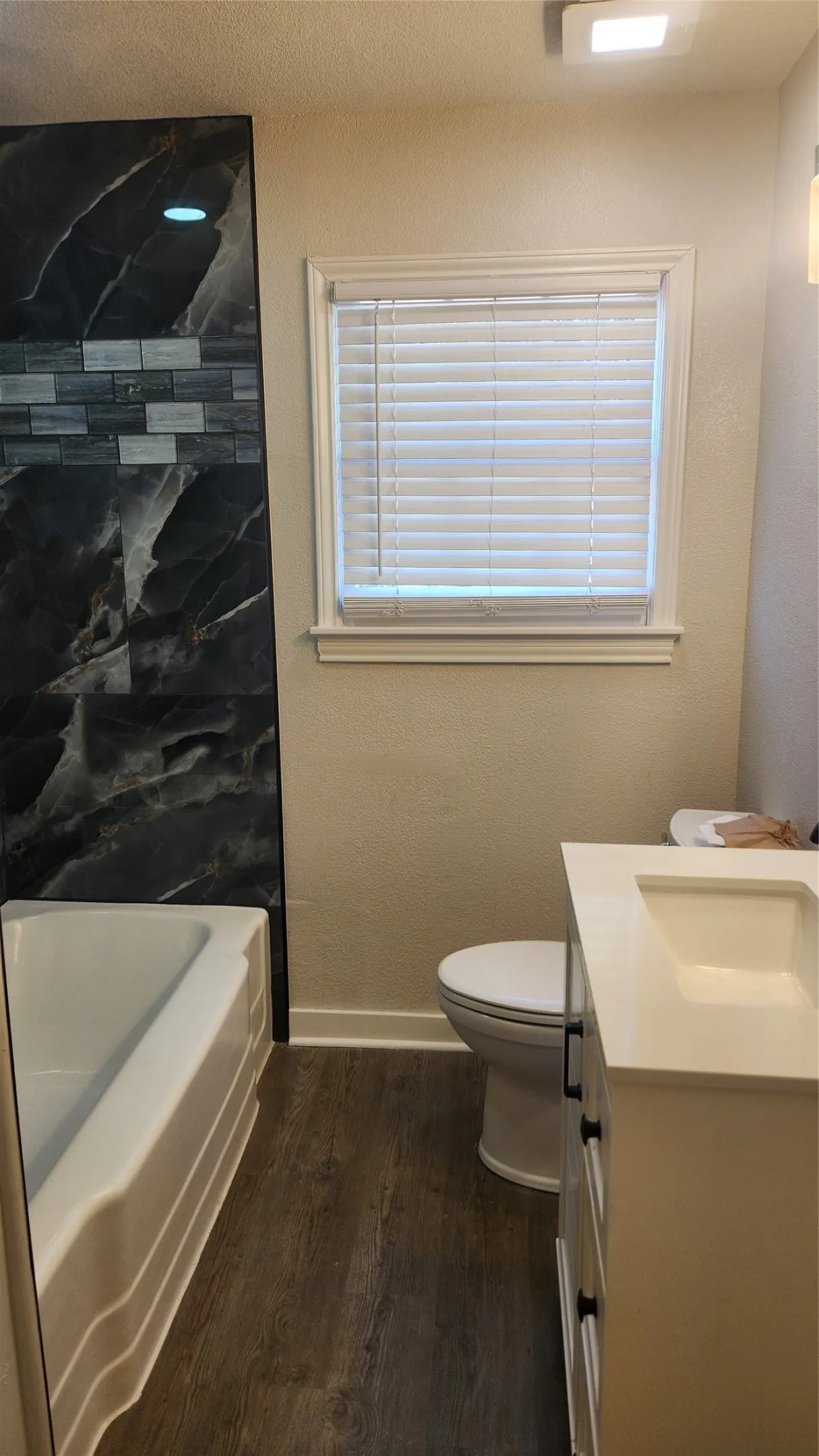 Bathtub with dark marble-look surround and decorative tile accent, wood-finish flooring, white vanity with integrated sink, and white toilet