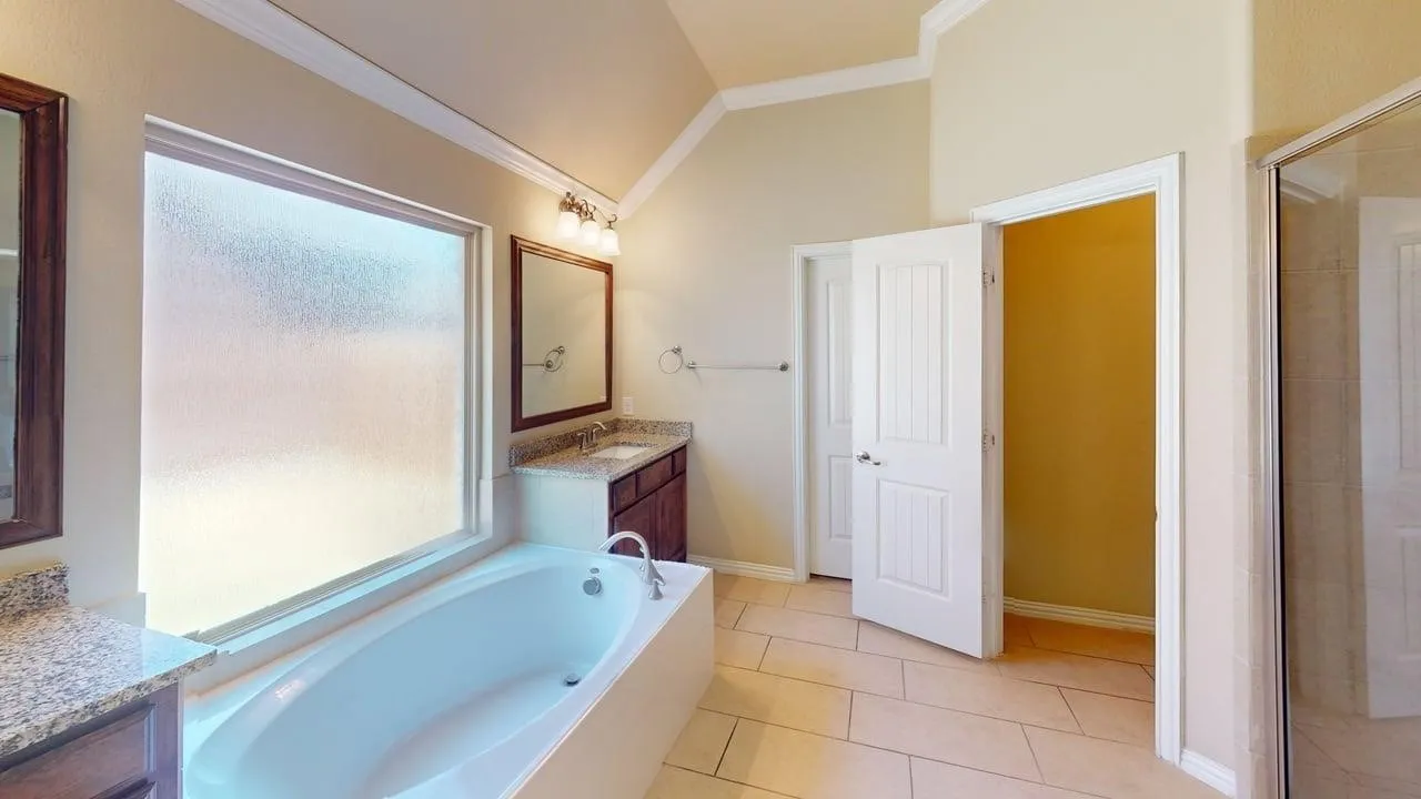 Soaking tub with a large privacy window, dual vanity sinks with stone countertops, and tile flooring