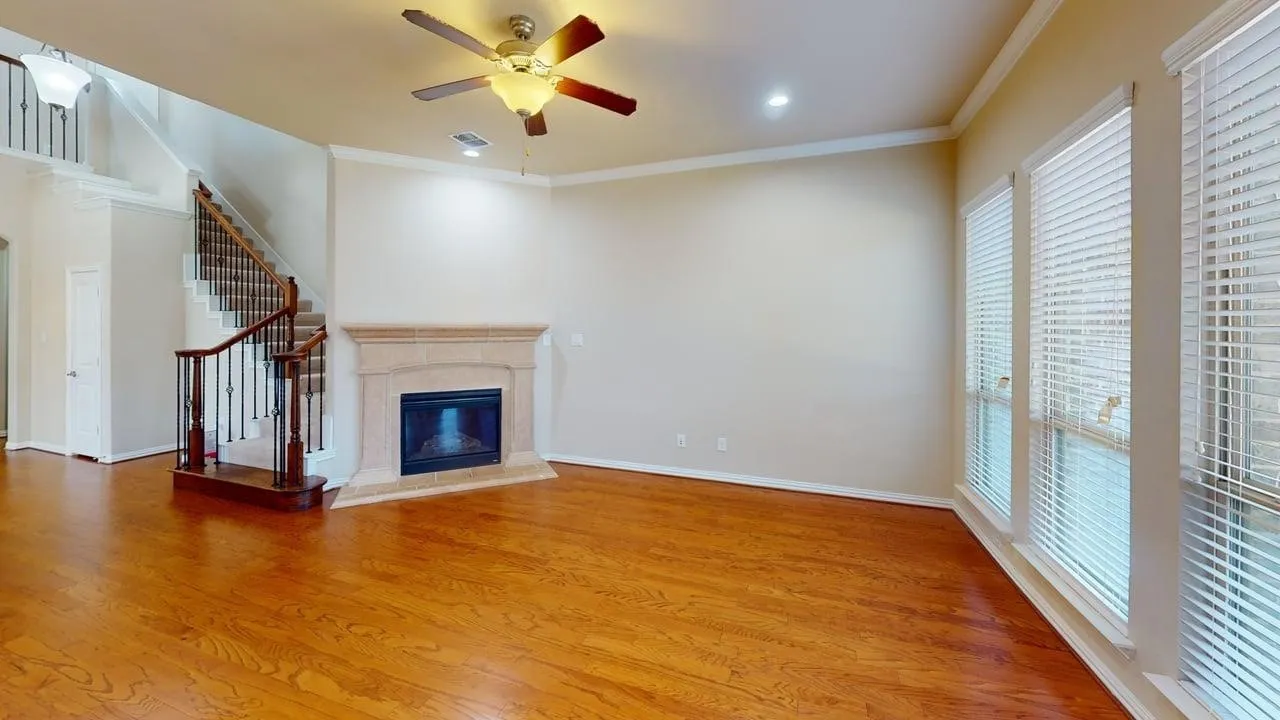 Wood-finish flooring, staircase with wrought iron balusters, stone-surround fireplace, ceiling fan with integrated lighting, and multiple windows with blinds