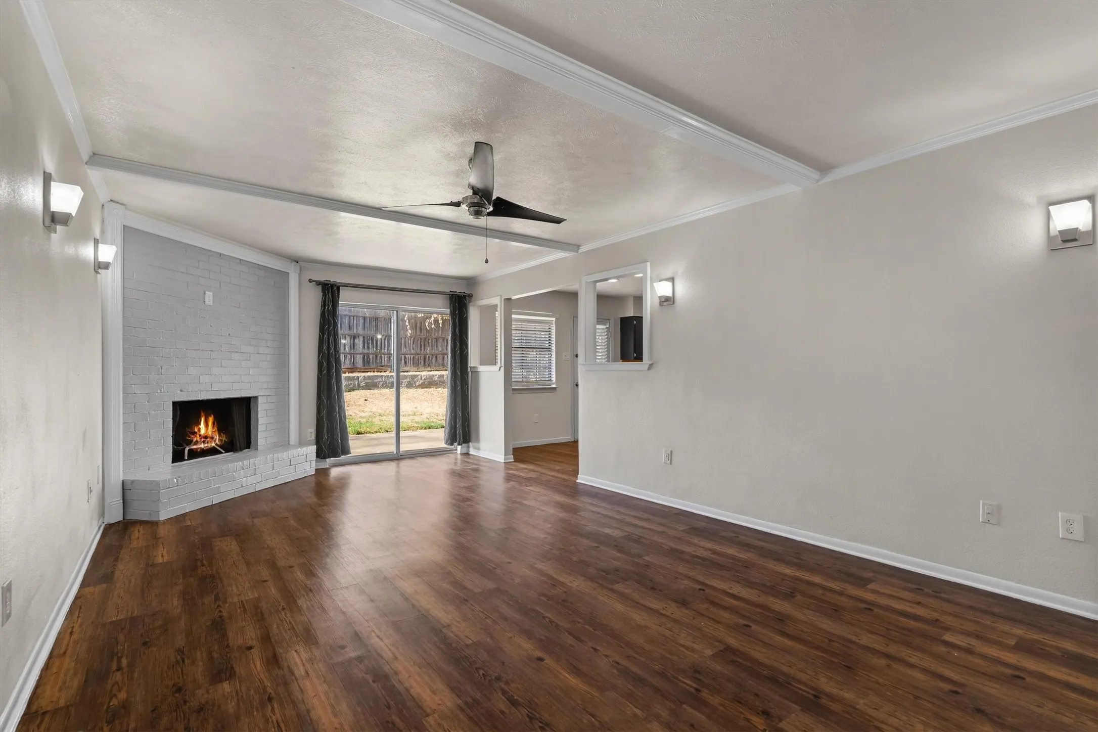 Wood-look flooring throughout, featuring a corner brick fireplace, exposed ceiling beams, and a ceiling fan