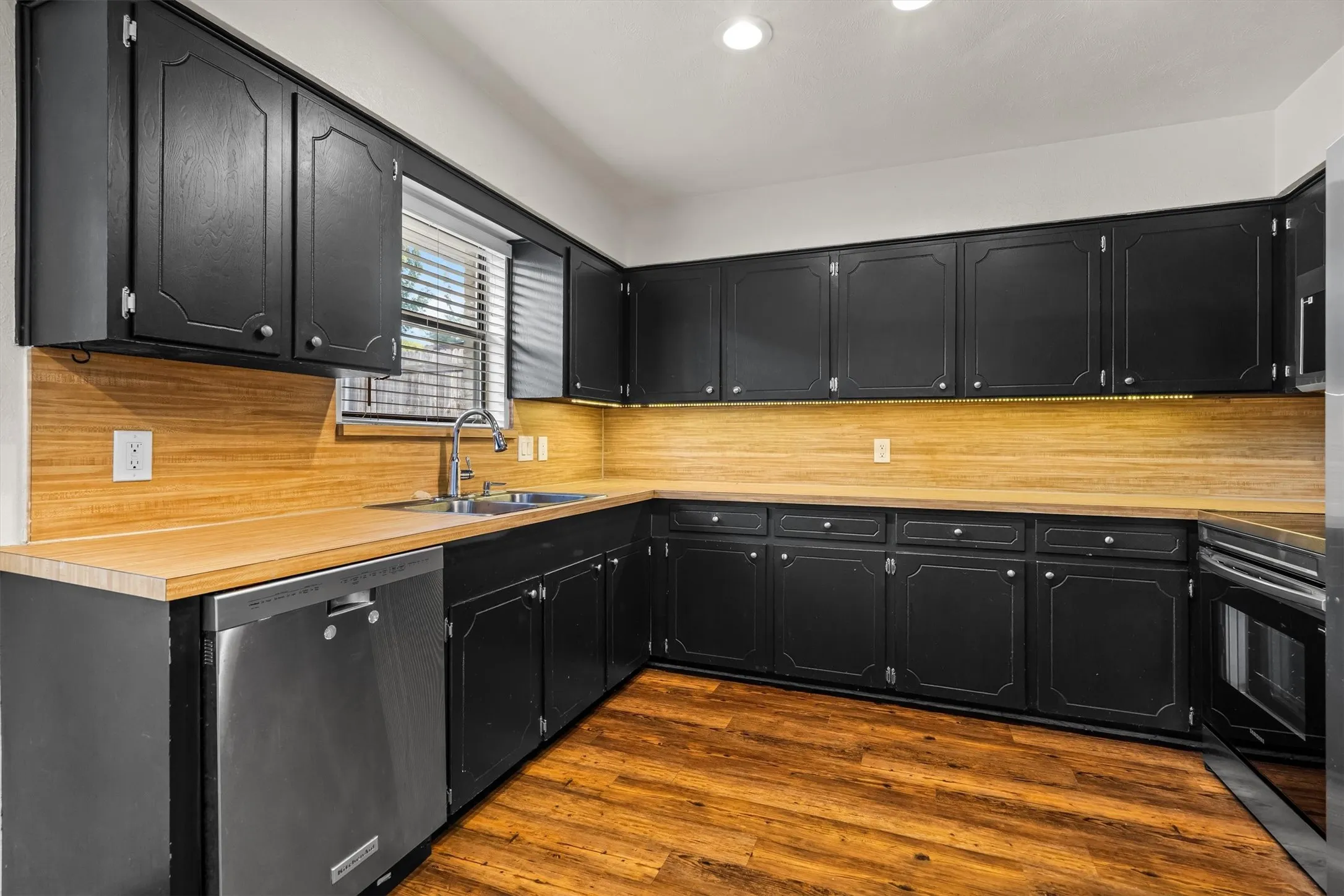 Kitchen featuring black cabinetry, wood-finish countertops, wood-finish flooring, and a stainless steel dishwasher