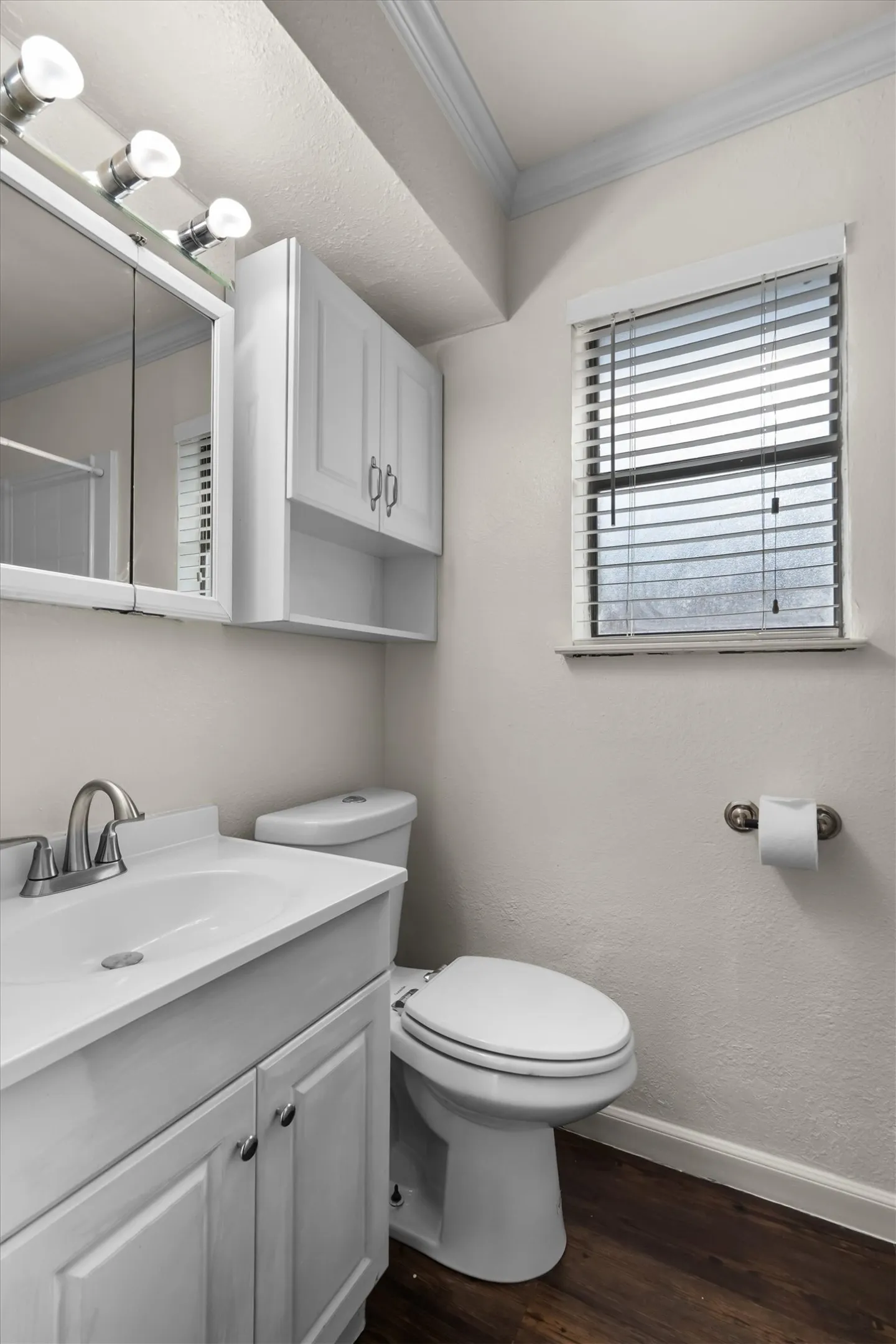 White vanity with integrated sink, brushed nickel faucet, wood-finish flooring, white wall cabinets, and a window with blinds