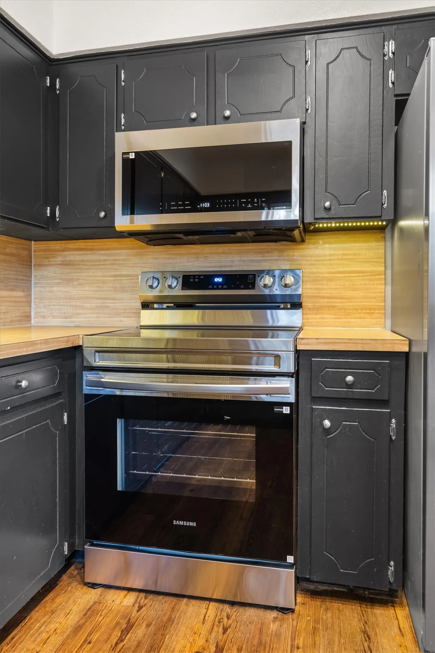 Black cabinetry, wood-look countertops, stainless steel appliances, wood-finish flooring, and a coordinating backsplash