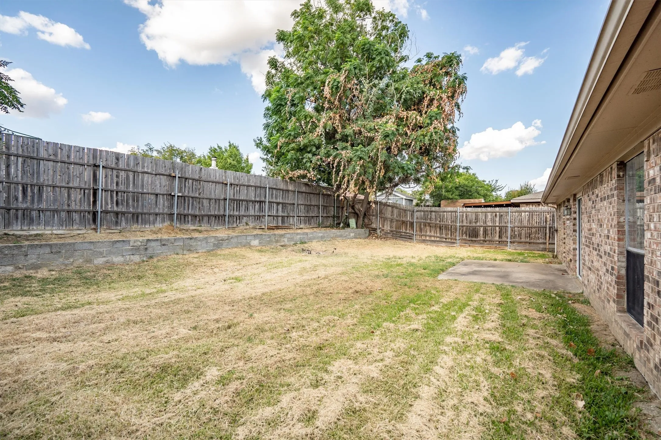 Expansive backyard with a full privacy fence, mature trees, a concrete patio, and a brick exterior wall