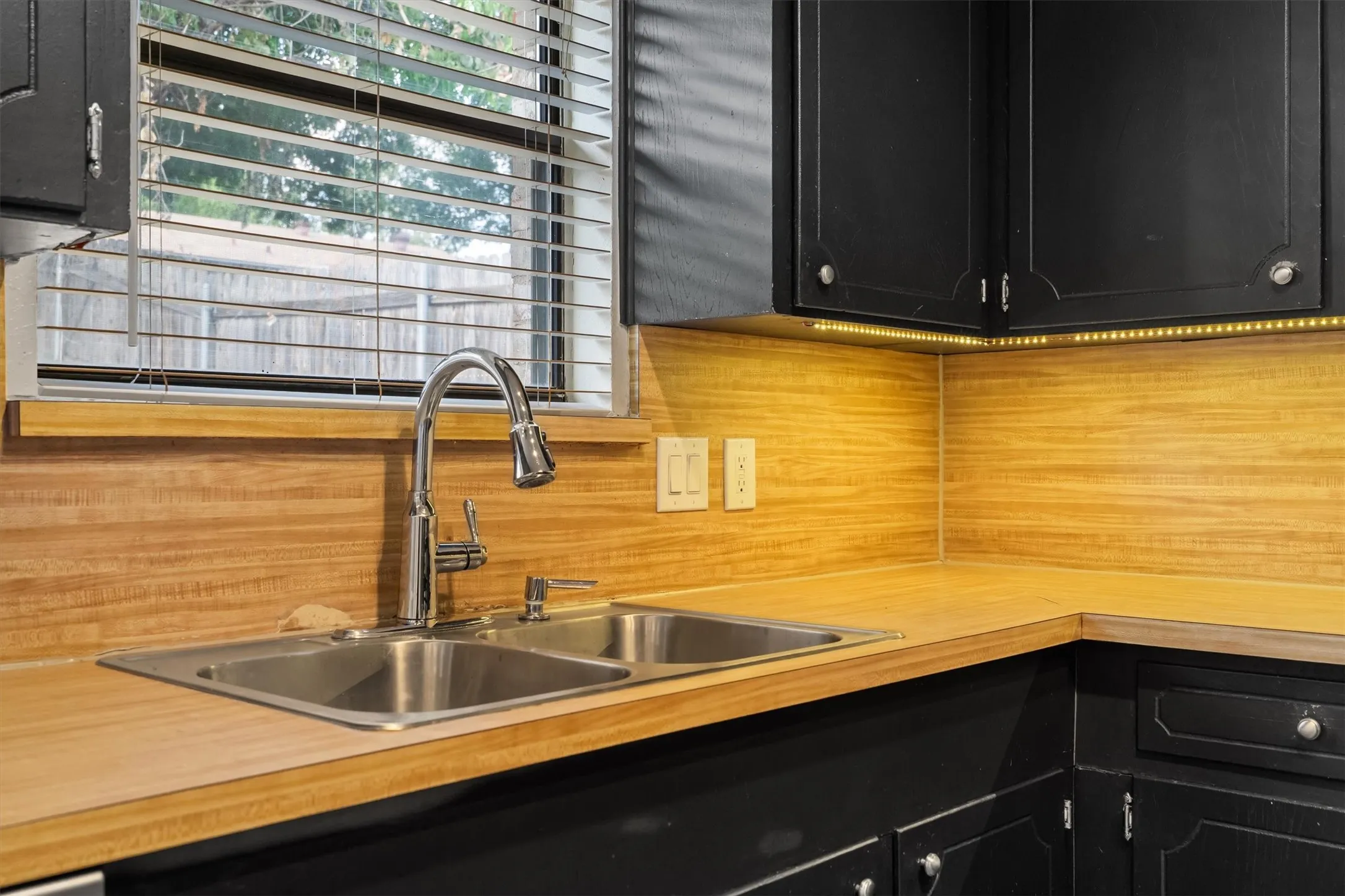 Dual basin stainless steel sink with pull-down faucet, wood-finish countertops and backsplash, black painted cabinetry, and under-cabinet lighting
