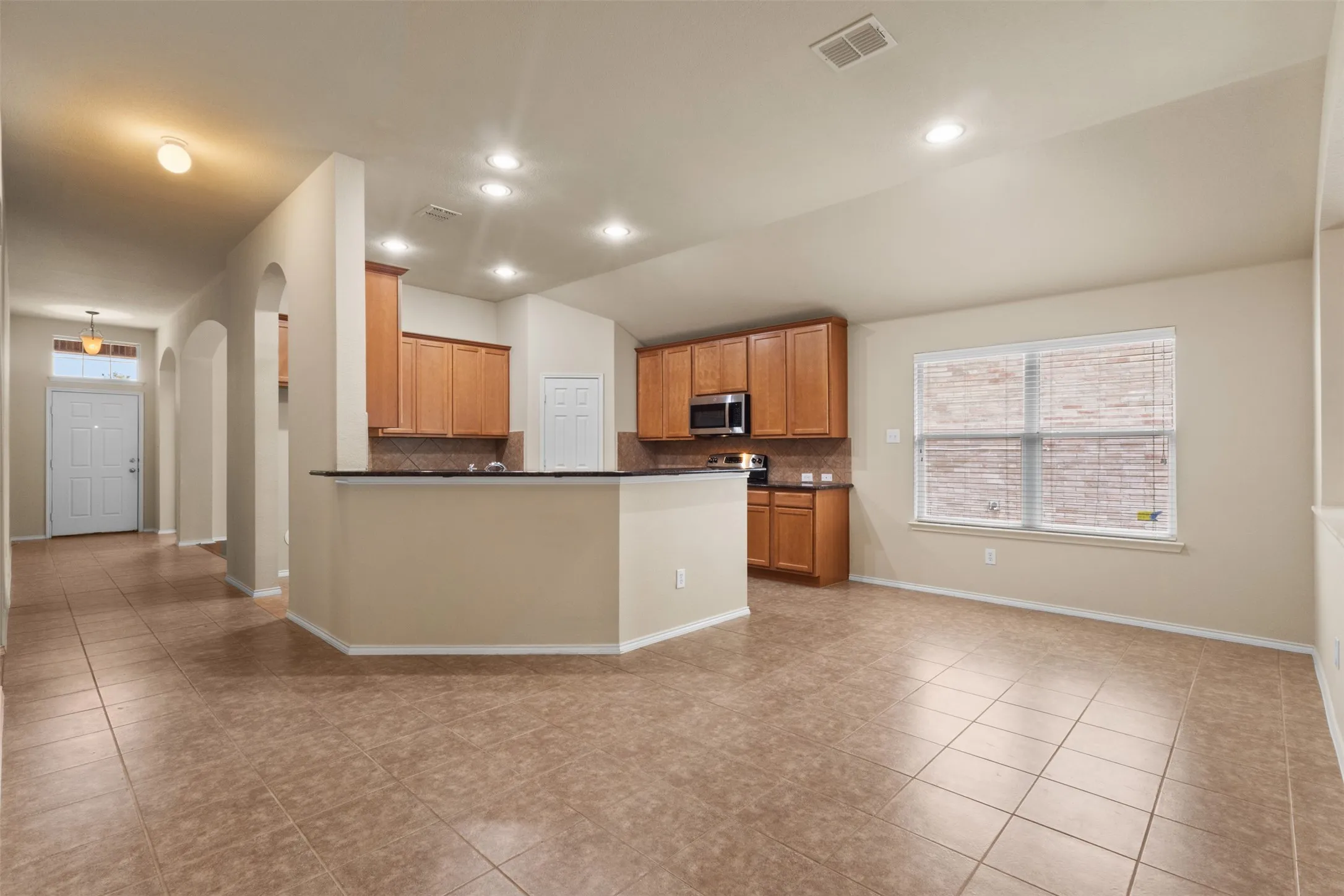 Open-concept living and kitchen area featuring wood-finish cabinetry, stainless steel appliances, tile flooring throughout, and abundant natural light from large windows