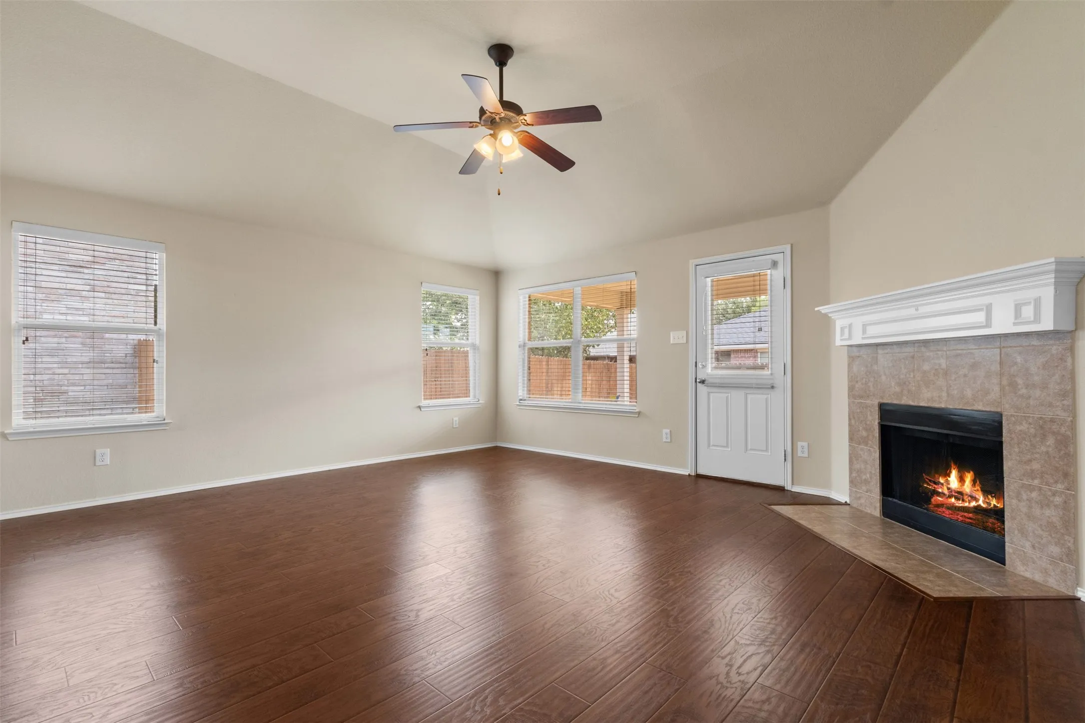 Vaulted ceiling with ceiling fan, wood-finish flooring, multiple windows, tiled fireplace with white mantel, and rear door access