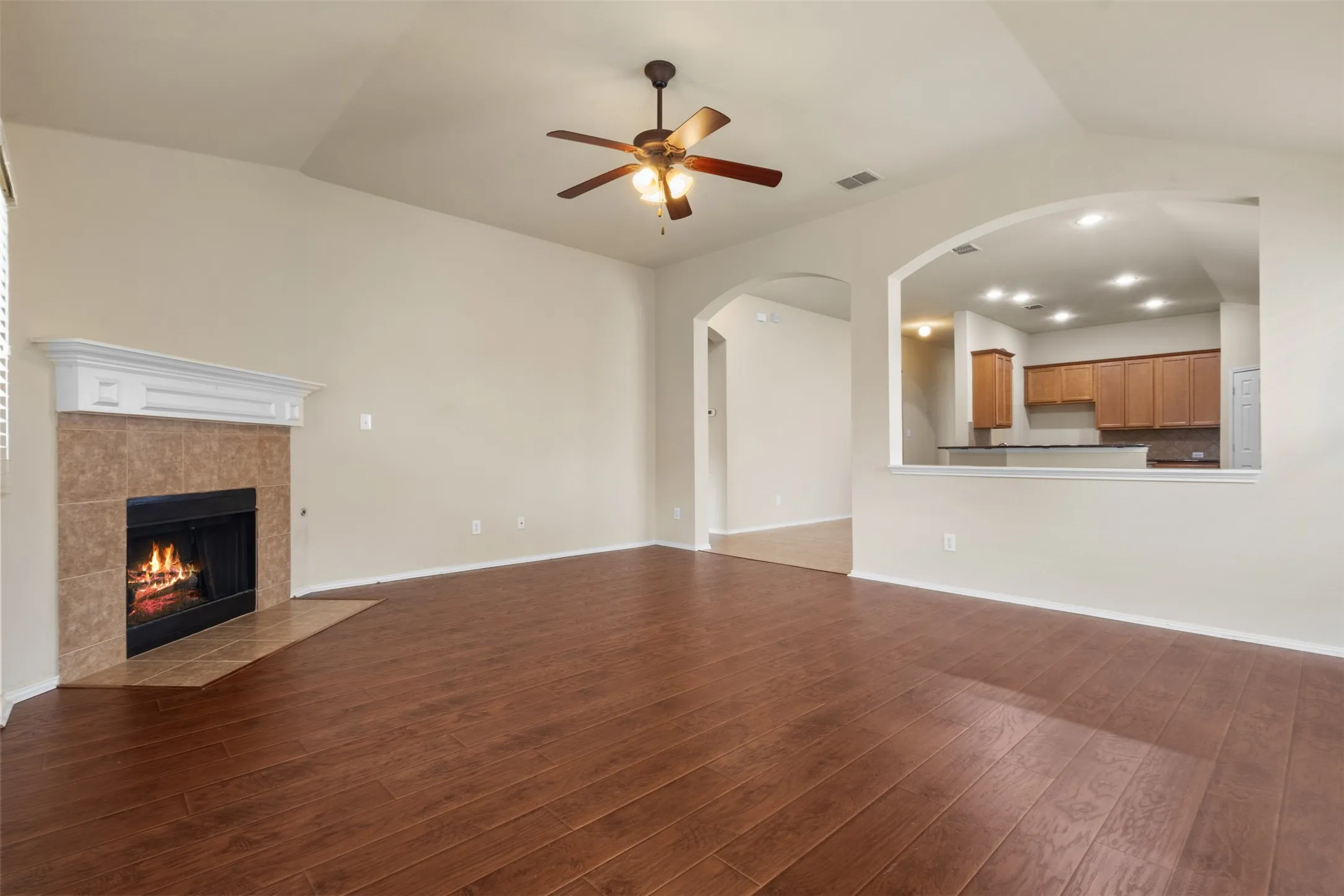Vaulted ceiling with a ceiling fan, wood-finish flooring, tiled fireplace with a mantel, and an arched pass-through to the kitchen featuring wood cabinetry