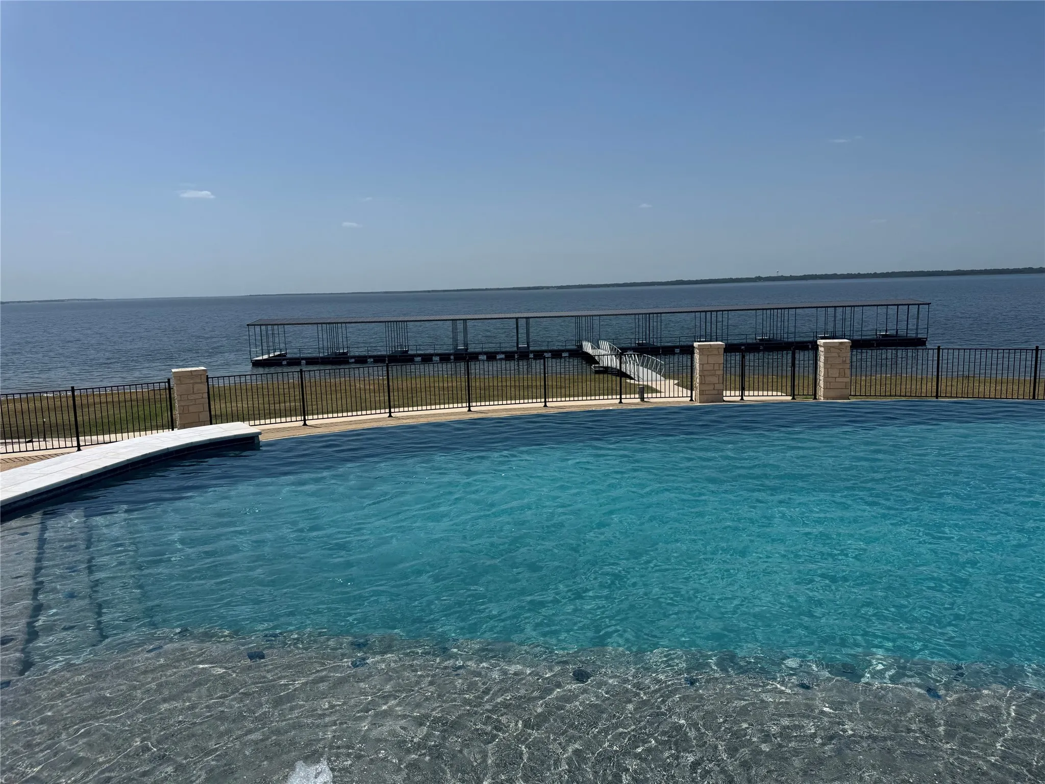 Infinity-edge swimming pool with a black metal fence featuring stone pillars, a private boat dock, and direct water frontage