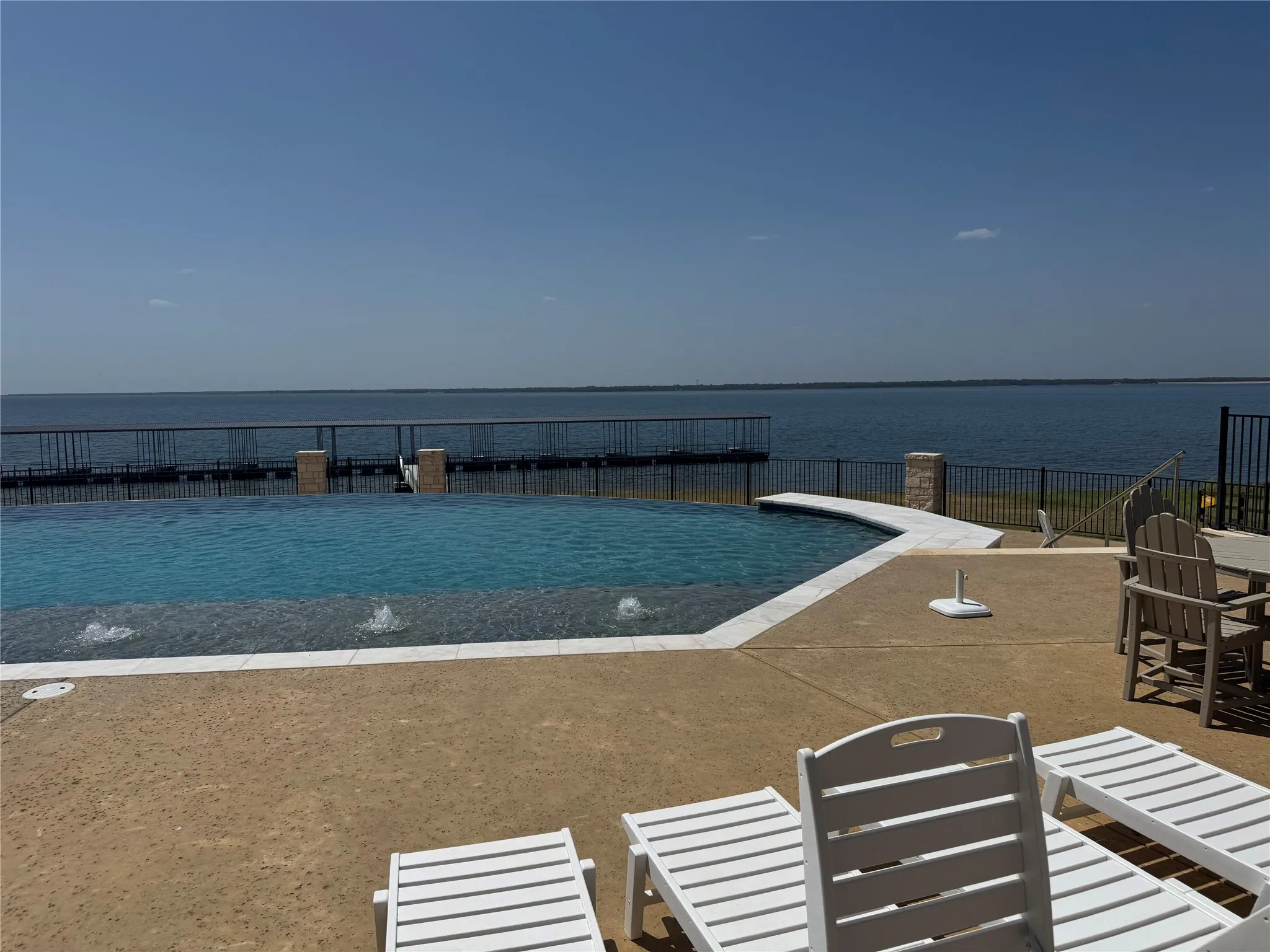 Infinity-edge pool with water features, expansive concrete patio, and a black metal fence bordering the waterfront