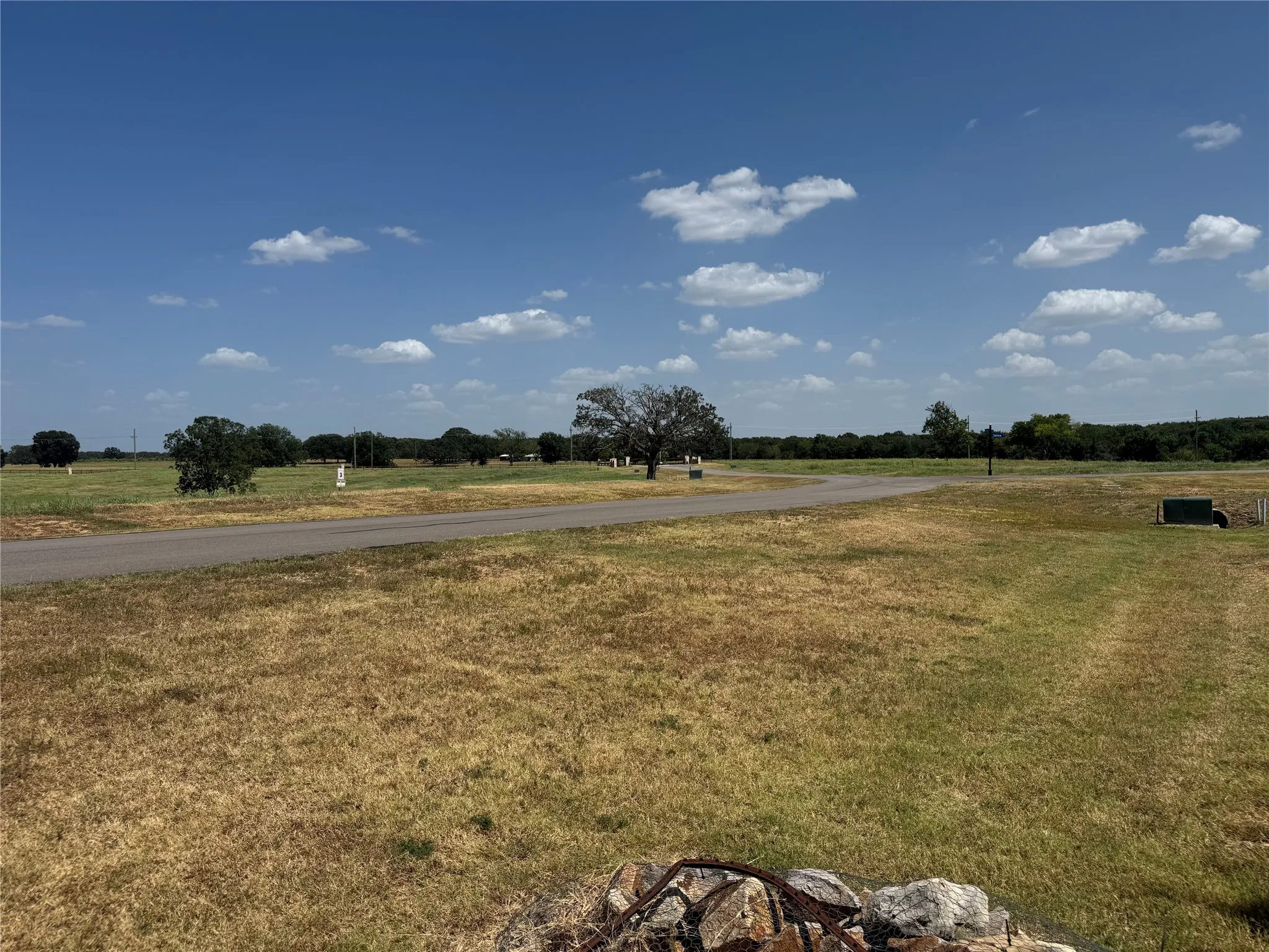 Expansive undeveloped land featuring a paved road, mature trees, and a stone-clad border