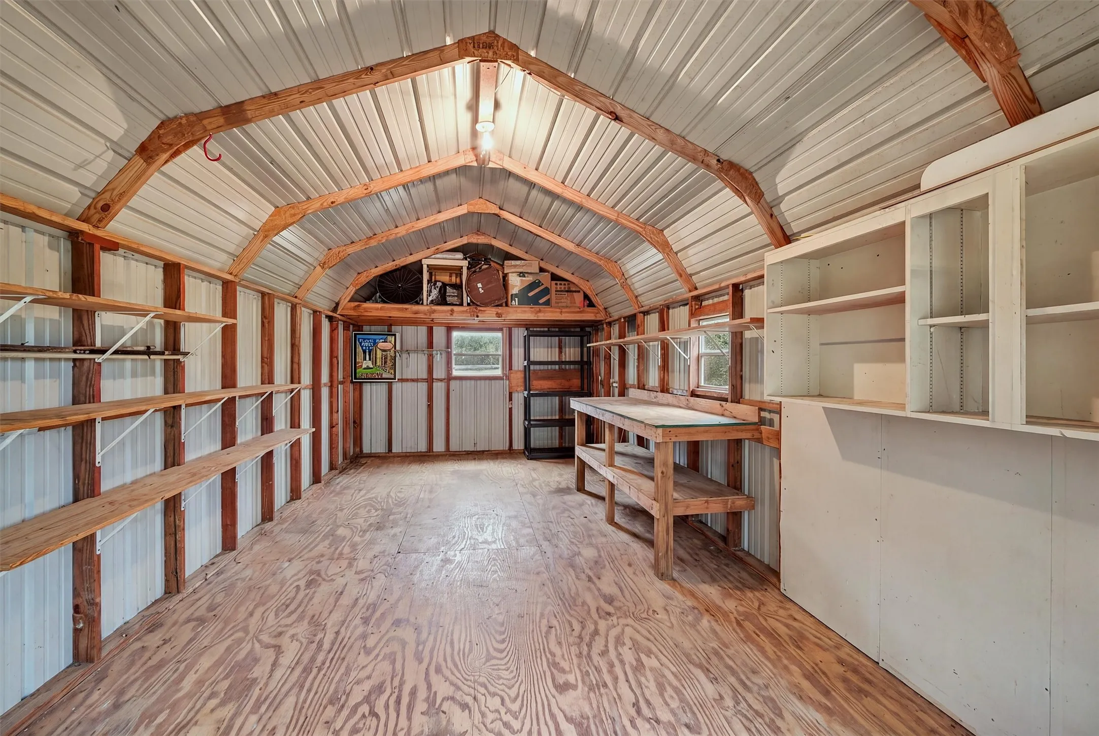 Interior of the multi-use storage shed with gabled metal roof, exposed wood beam construction, integrated shelving units, built-in workbench, and overhead cabinetry