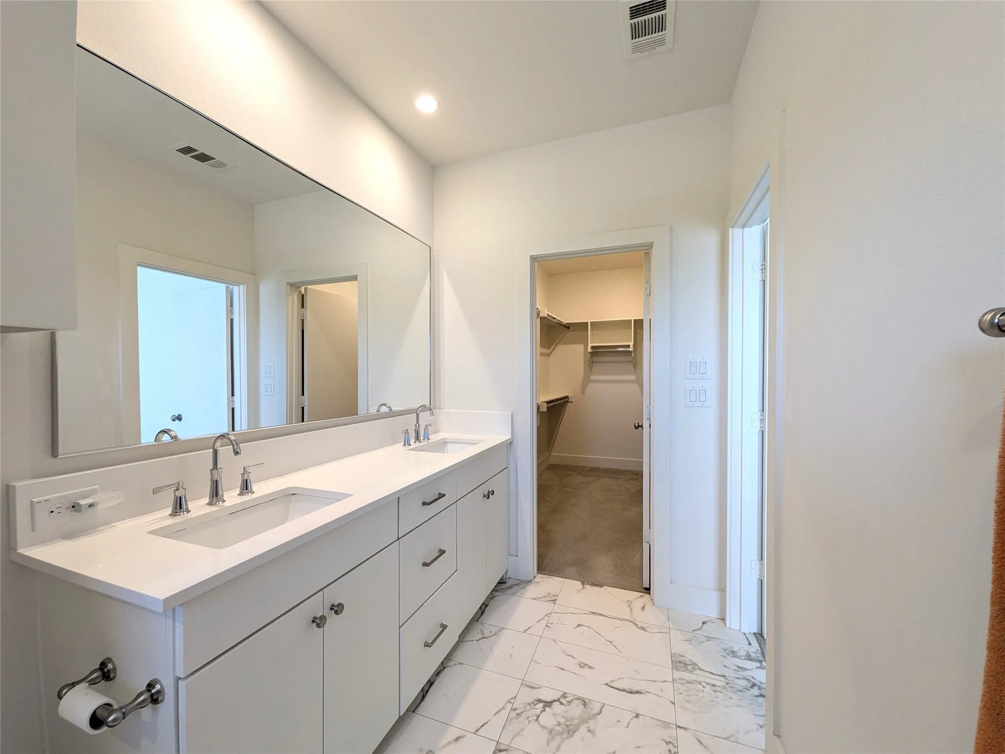 Dual vanity with white cabinetry, polished chrome fixtures, integrated sinks, large wall-mounted mirror, and marble-look tile flooring
