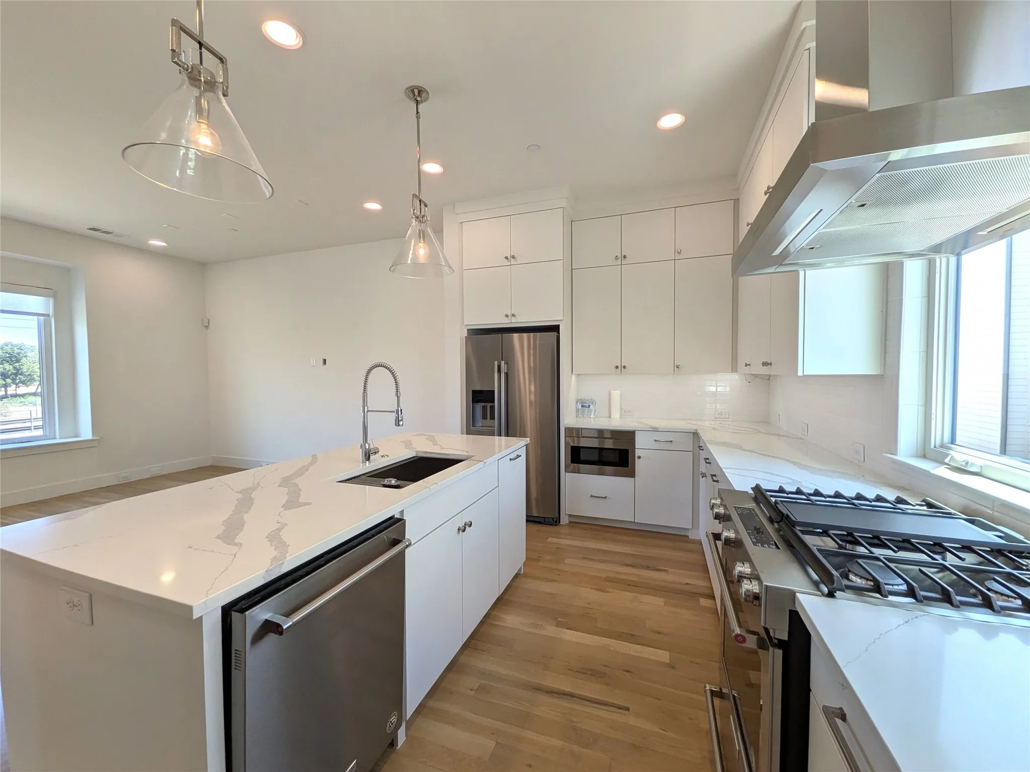 Chef's kitchen featuring white cabinetry with brushed nickel hardware, quartz countertops, a large waterfall-edge island with integrated sink and dishwasher, professional-grade gas cooktop with stainless steel range hood, stainless steel refrigerator, and abundant recessed and pendant lighting