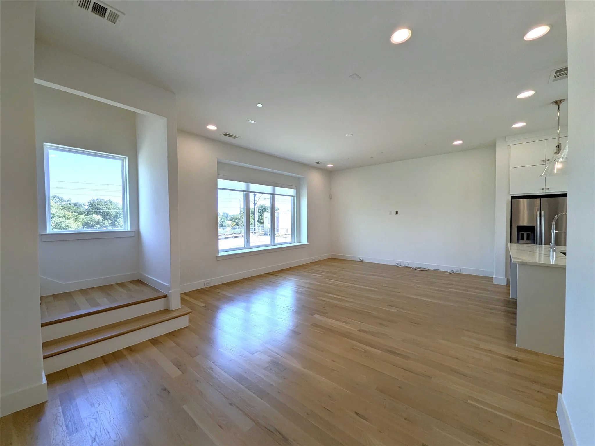 Bright, open living space featuring gleaming wood-finish flooring throughout, recessed ceiling lighting, and multiple large windows flooding the room with natural light