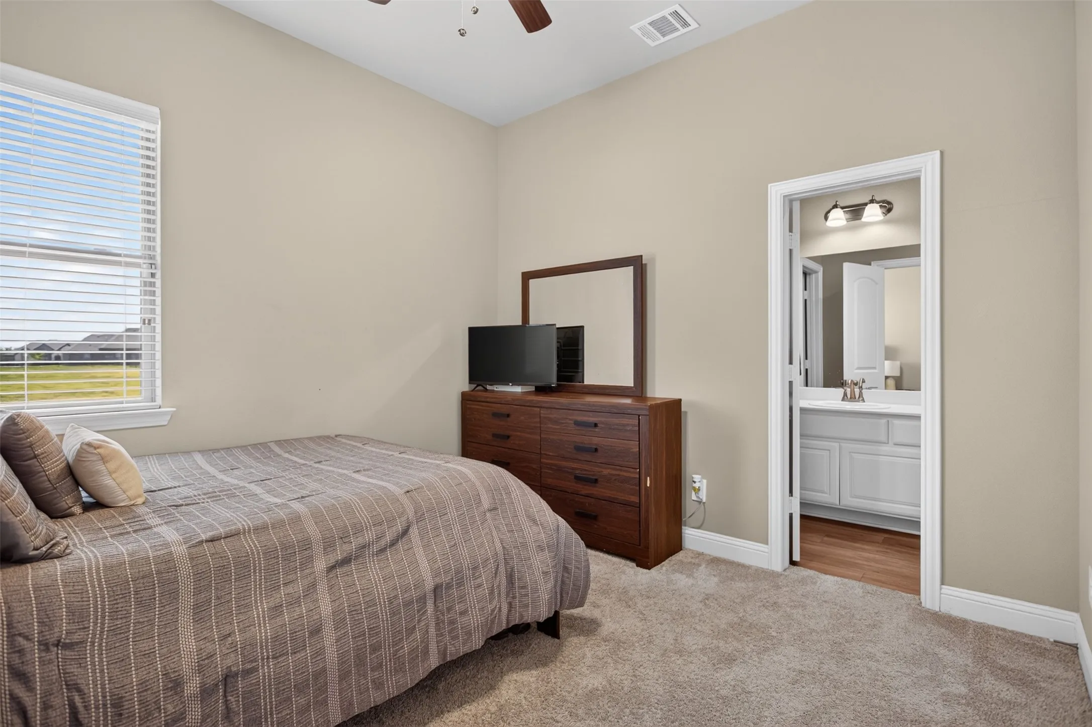 Vaulted ceiling, ceiling fan, window with blinds, carpeted flooring, and an attached bathroom with a vanity and wall-mounted light fixture
