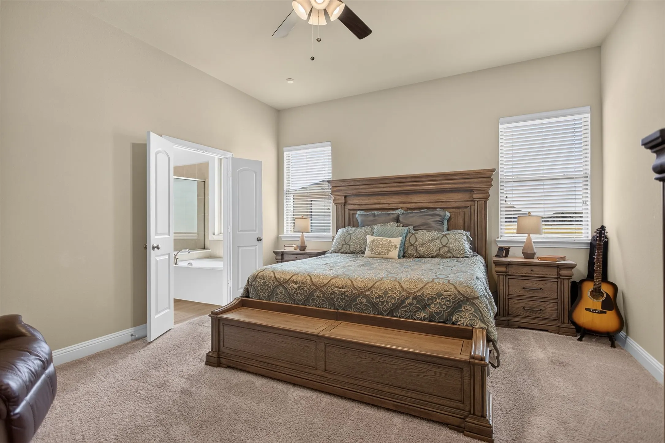 Carpeted bedroom with a ceiling fan, dual windows with blinds, and double doors opening to a bathroom featuring a built-in tub