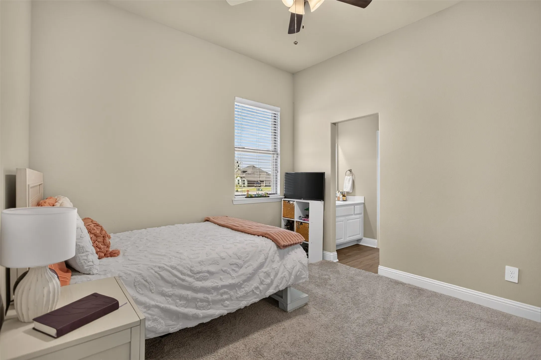 Carpeted flooring, overhead ceiling fan, window with blinds, and an entrance to an additional room with a vanity