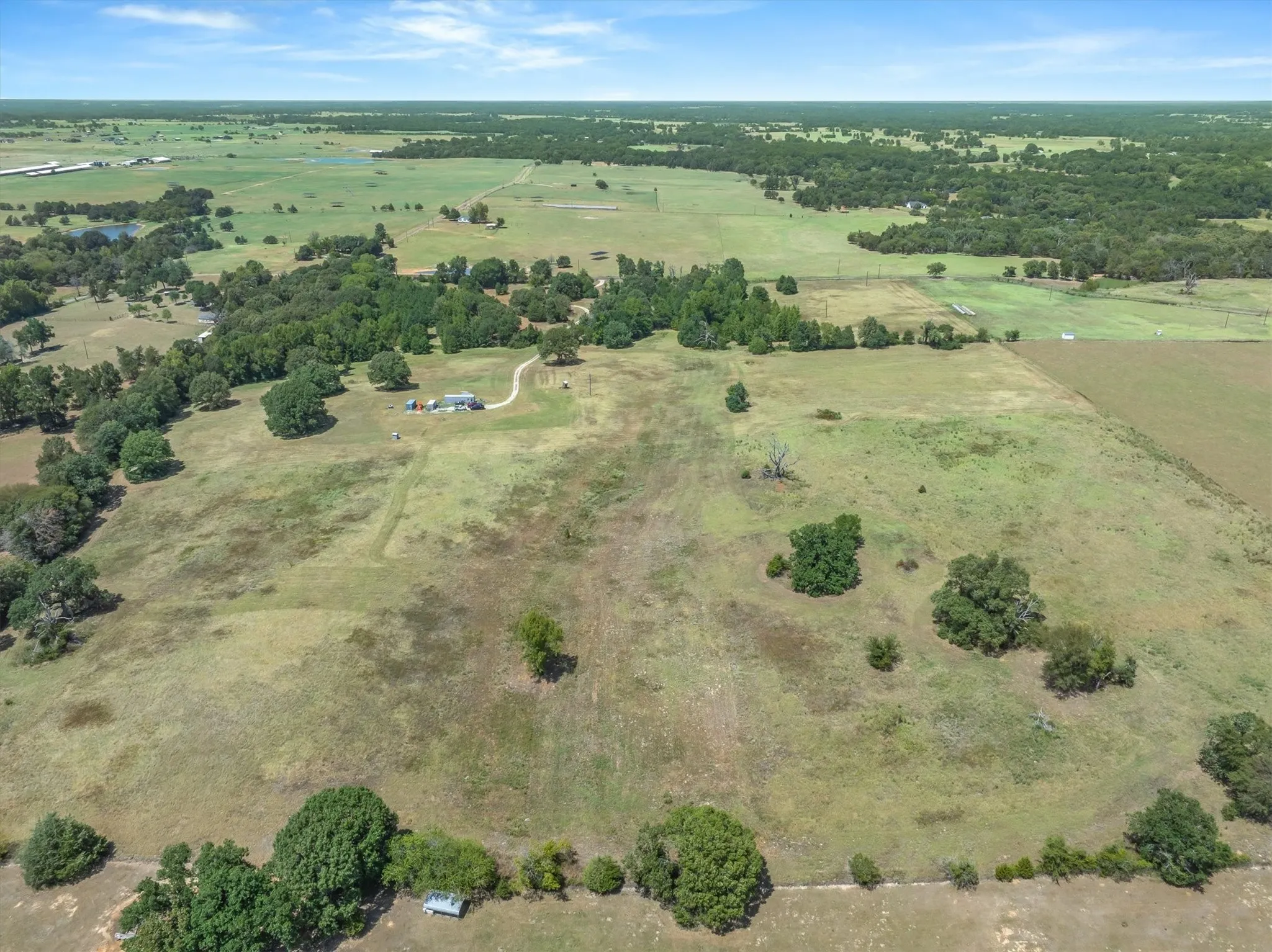 Expansive rural property featuring a single-story dwelling, large open fields, scattered mature trees, and a distant treeline