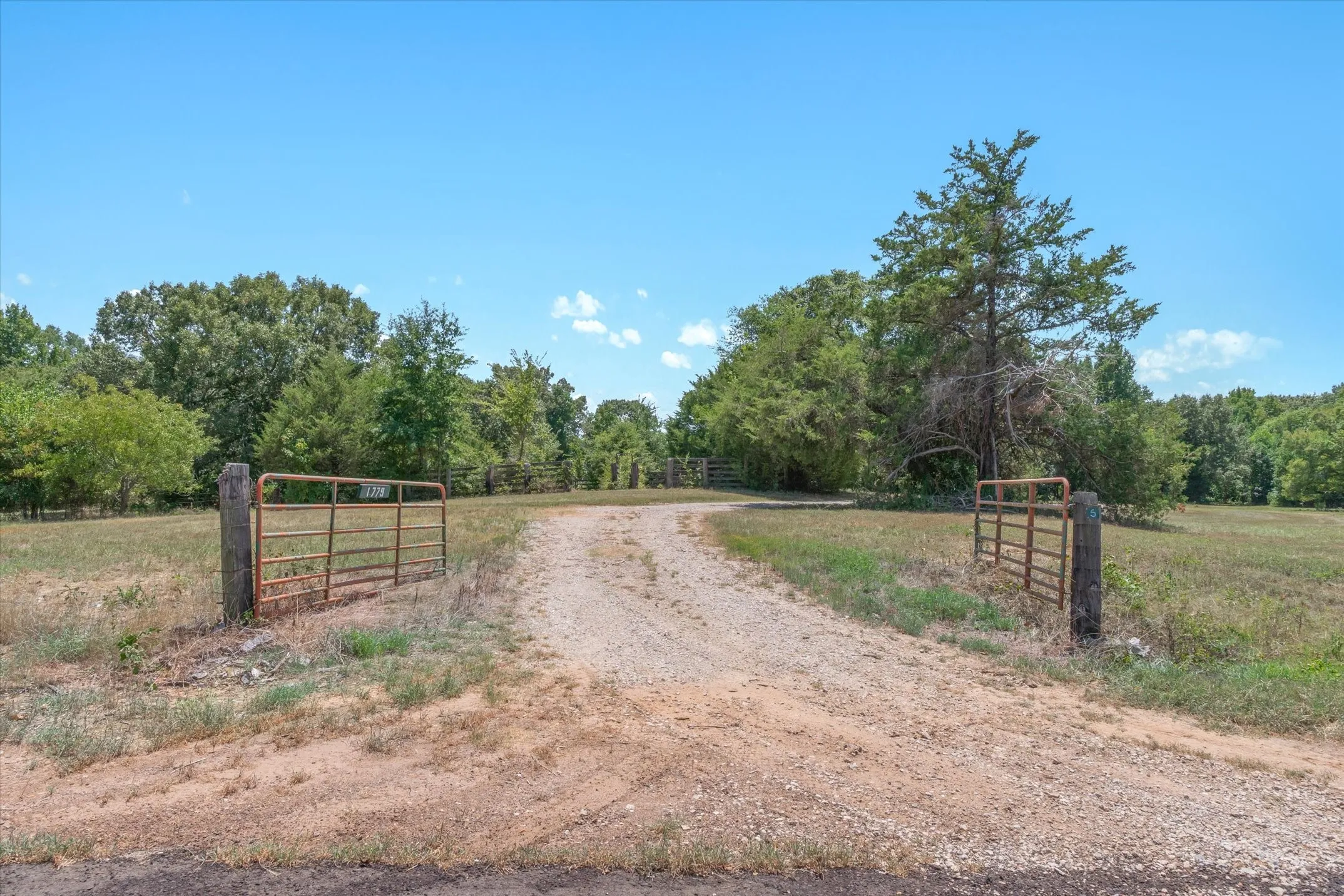 Gated gravel driveway leading to expansive land with mature trees and open fields