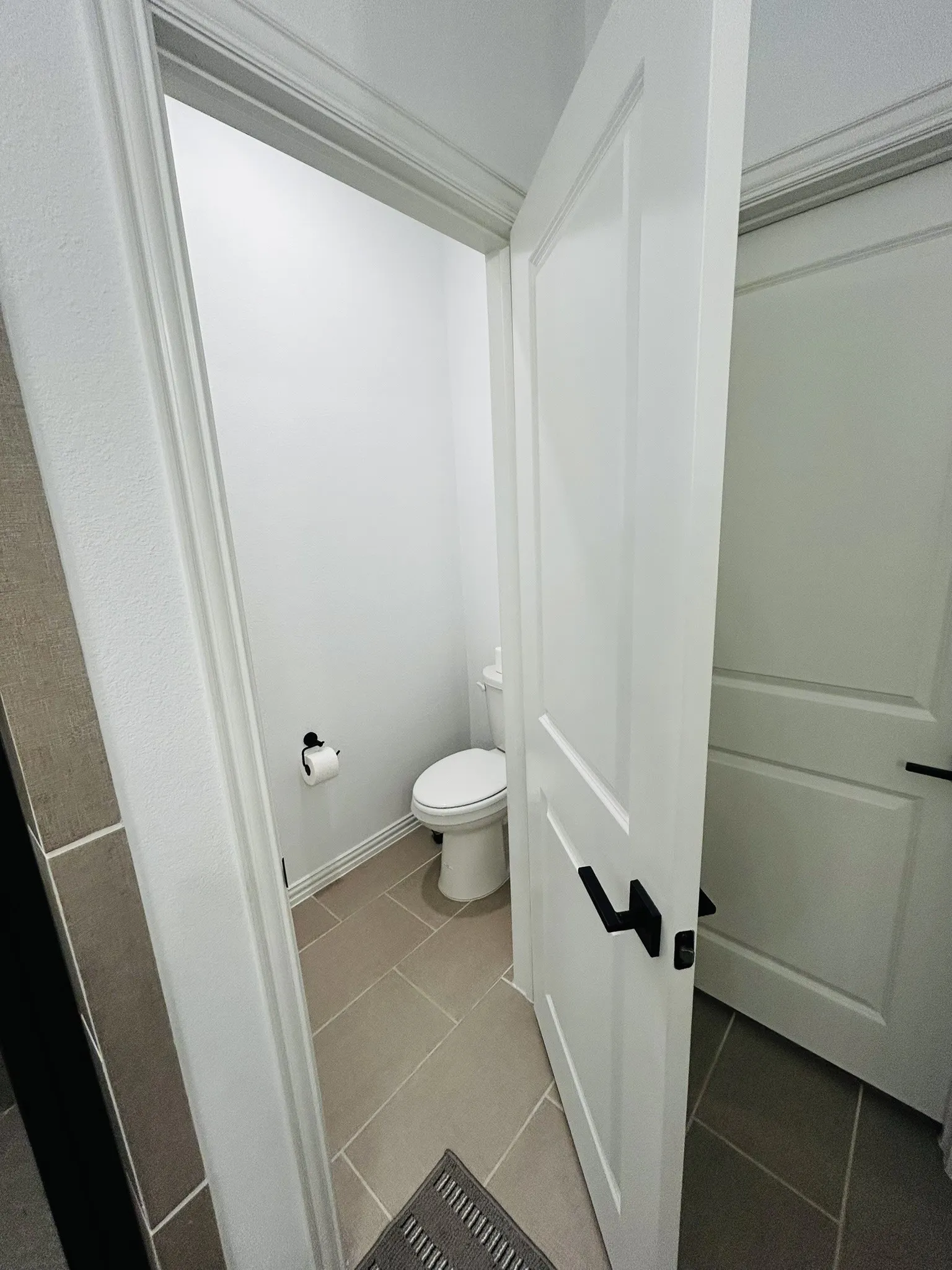 Private water closet with neutral-toned tile flooring, a solid core door with matte black hardware, and crisp white trim