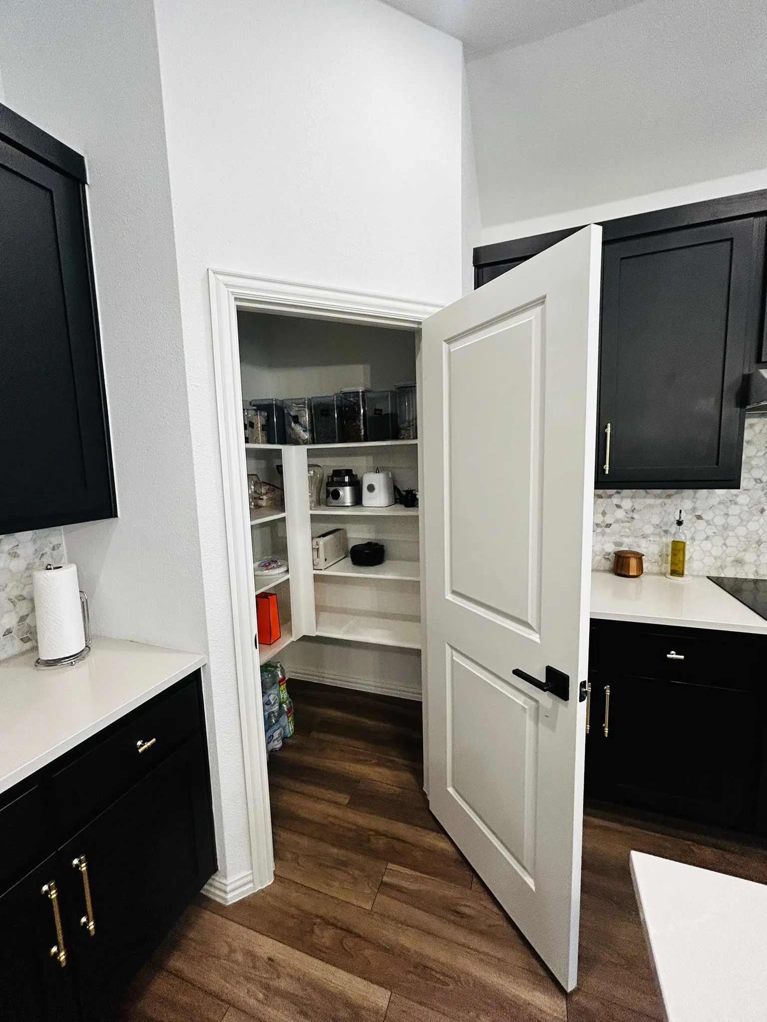 Walk-in pantry with built-in shelving, wood-finish flooring, white countertops, black cabinetry, and mosaic tile backsplash