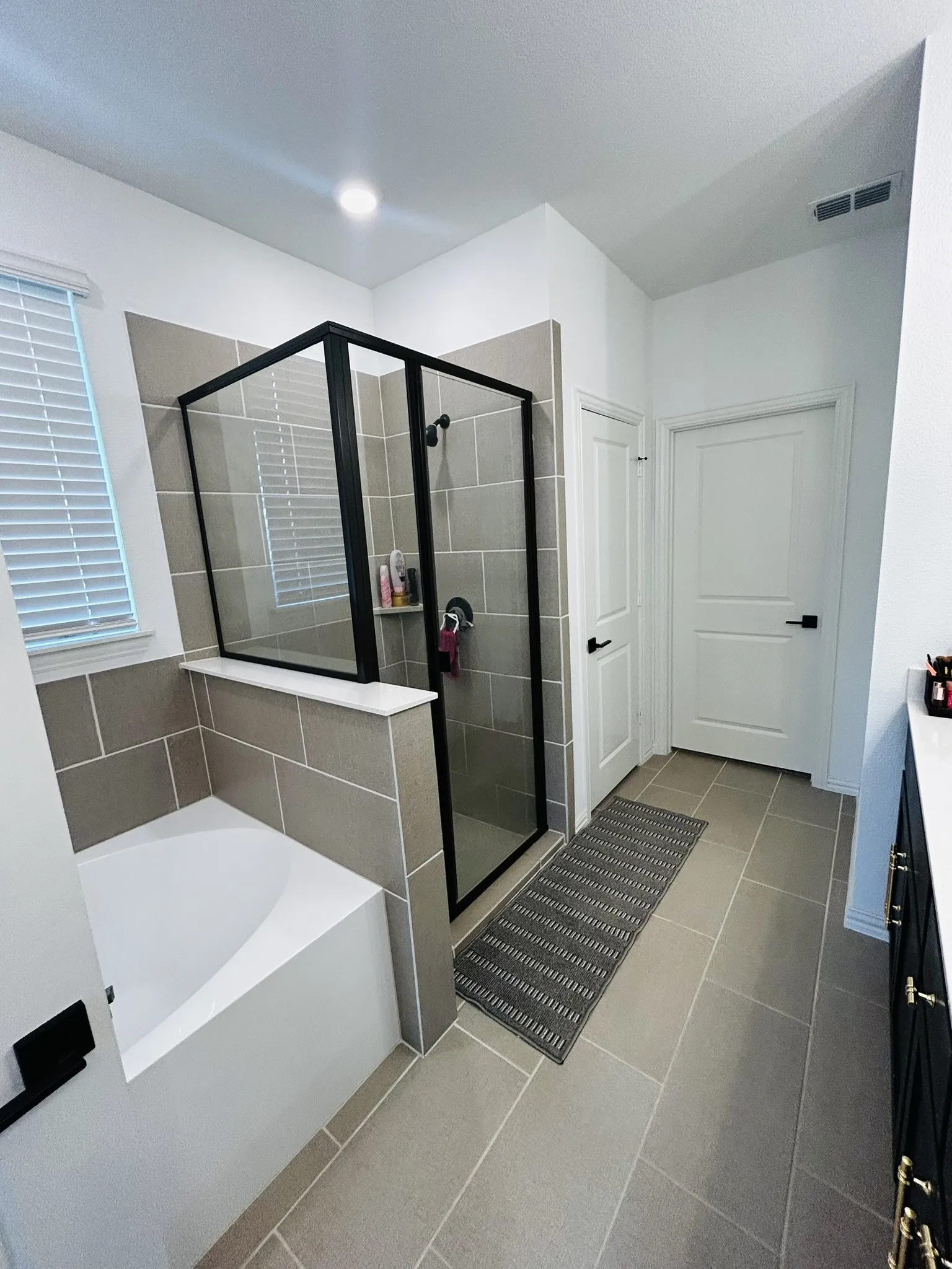 Tile flooring with white grout lines, a built-in bathtub, a walk-in shower with black-framed glass enclosure, and a vanity with black cabinetry and gold-toned hardware