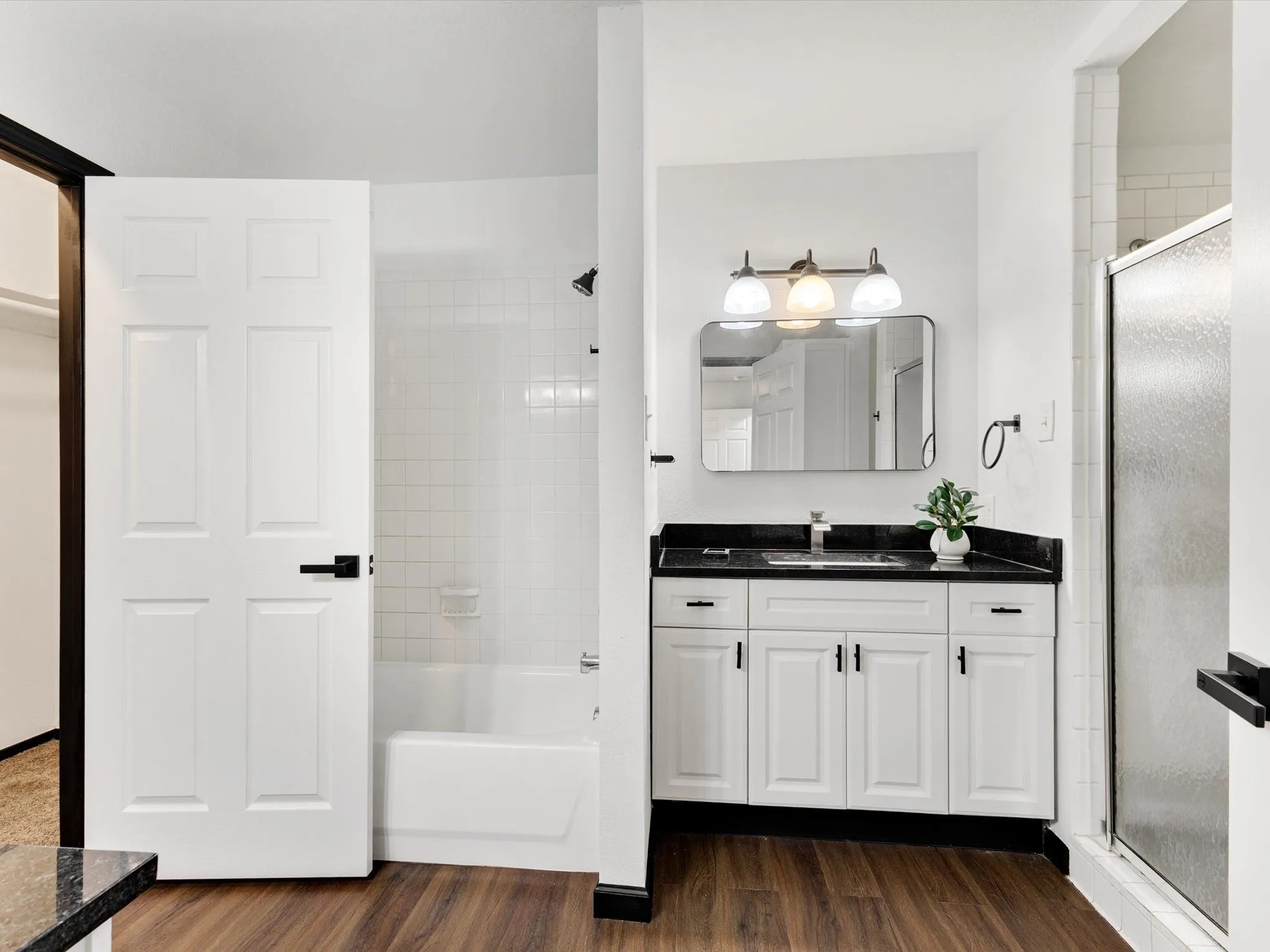 White paneled door with black hardware, white subway tile shower surround, wood-finish flooring, white vanity with black countertop, and a rectangular mirror with a three-light fixture