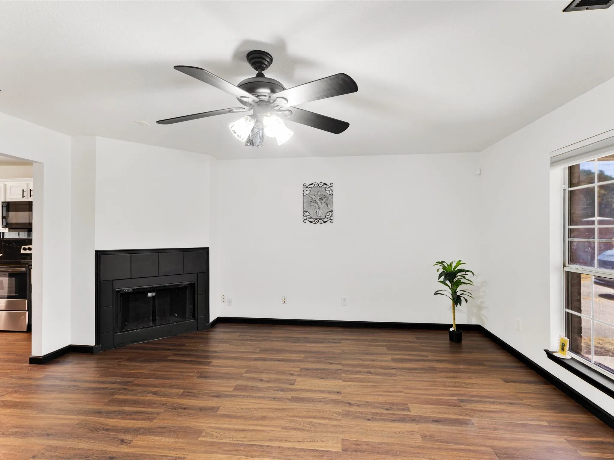 Wood-finish flooring throughout, featuring a corner fireplace with a black mantel, a ceiling fan with integrated lighting, and a large window providing natural light